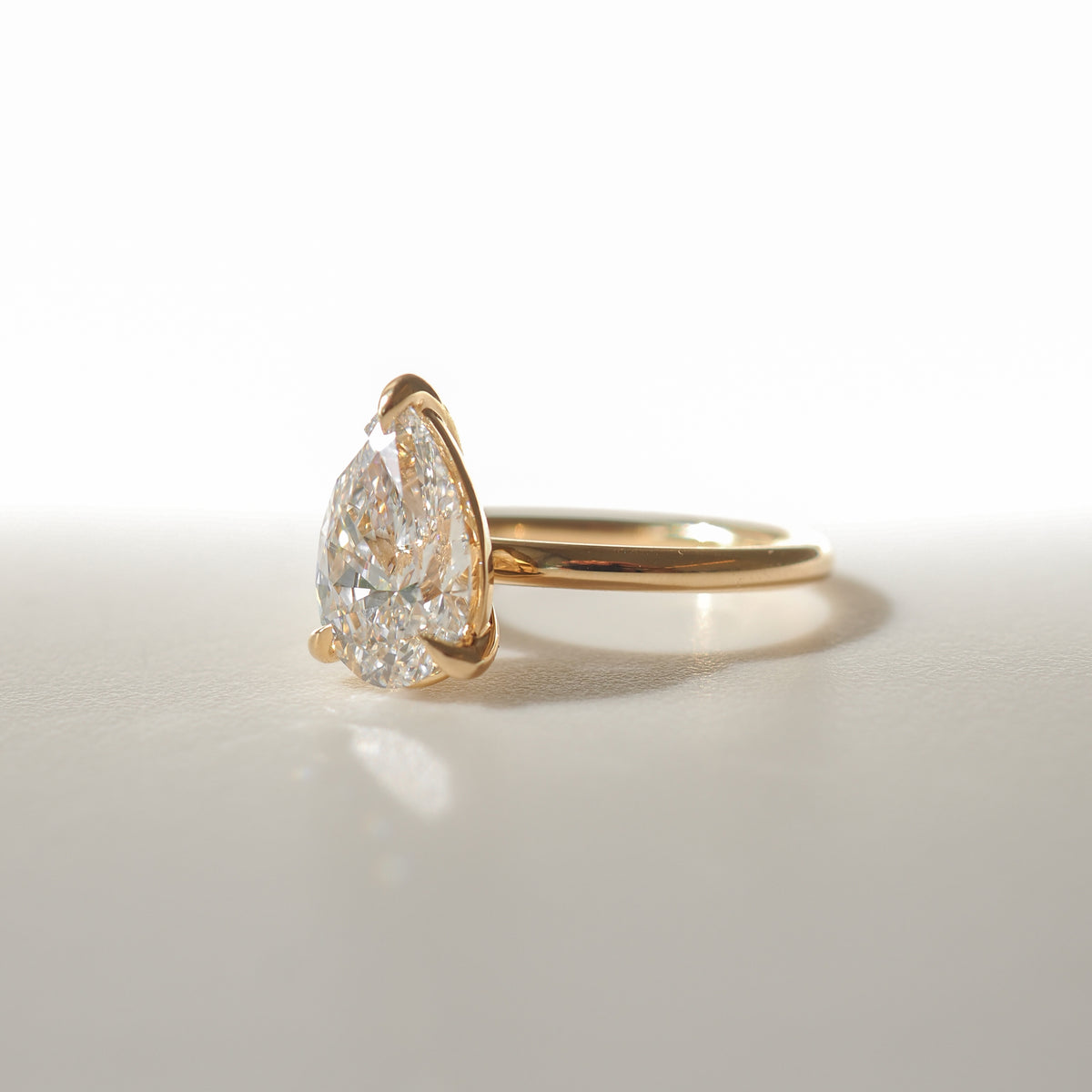 Gold ring with large pear-shaped diamond, elegantly set on a sleek band, reflecting light on a white surface. Luxurious jewellery piece.