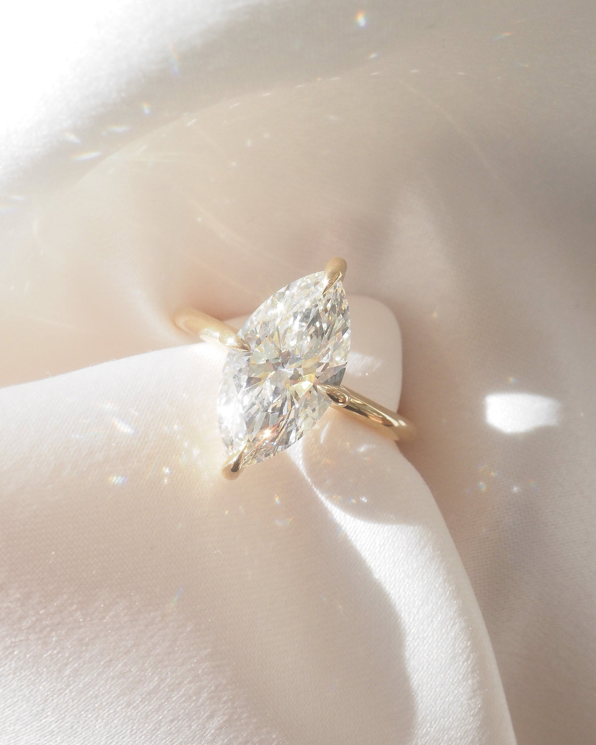 A yellow gold ring with a marquise-cut clear gemstone, resting on draped white satin fabric.