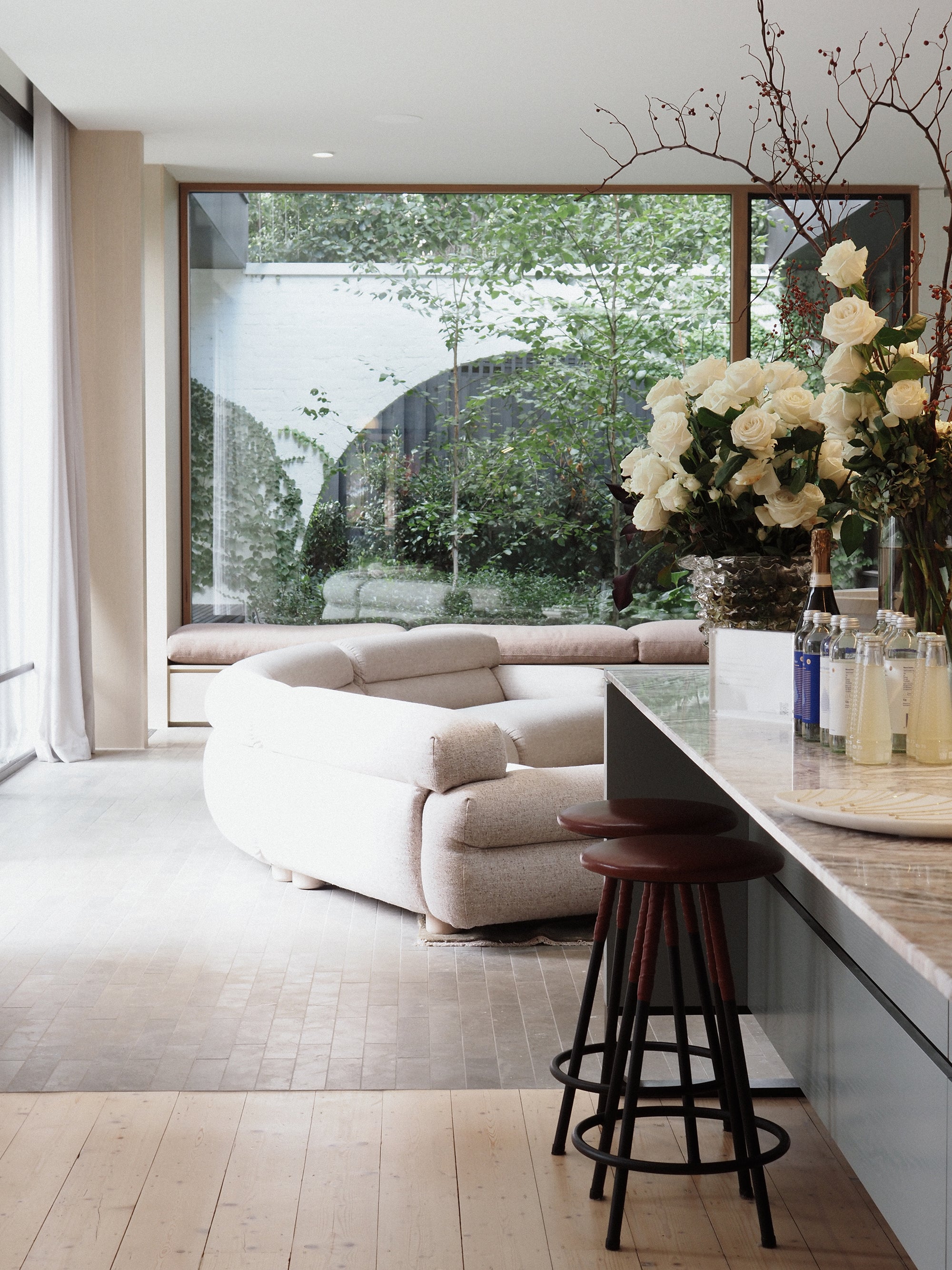 Luxurious modern interior with plush sofa, marble counter, elegant bar stools, and tall windows showcasing a lush garden. Styled with white roses.