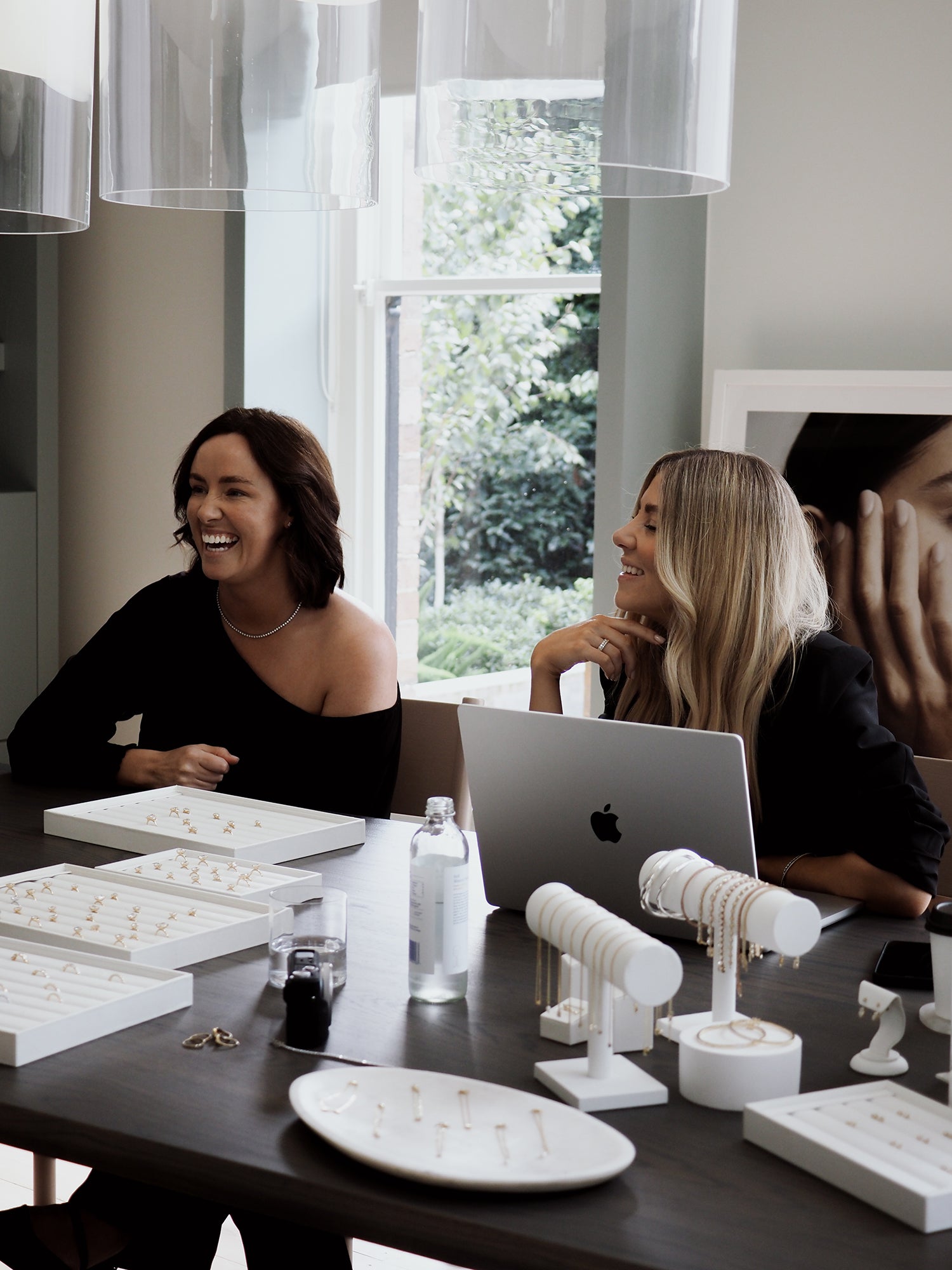 Two women sit at a table displaying luxury jewellery, with laptops and trays of gold pieces. The setting is elegant and well-lit.