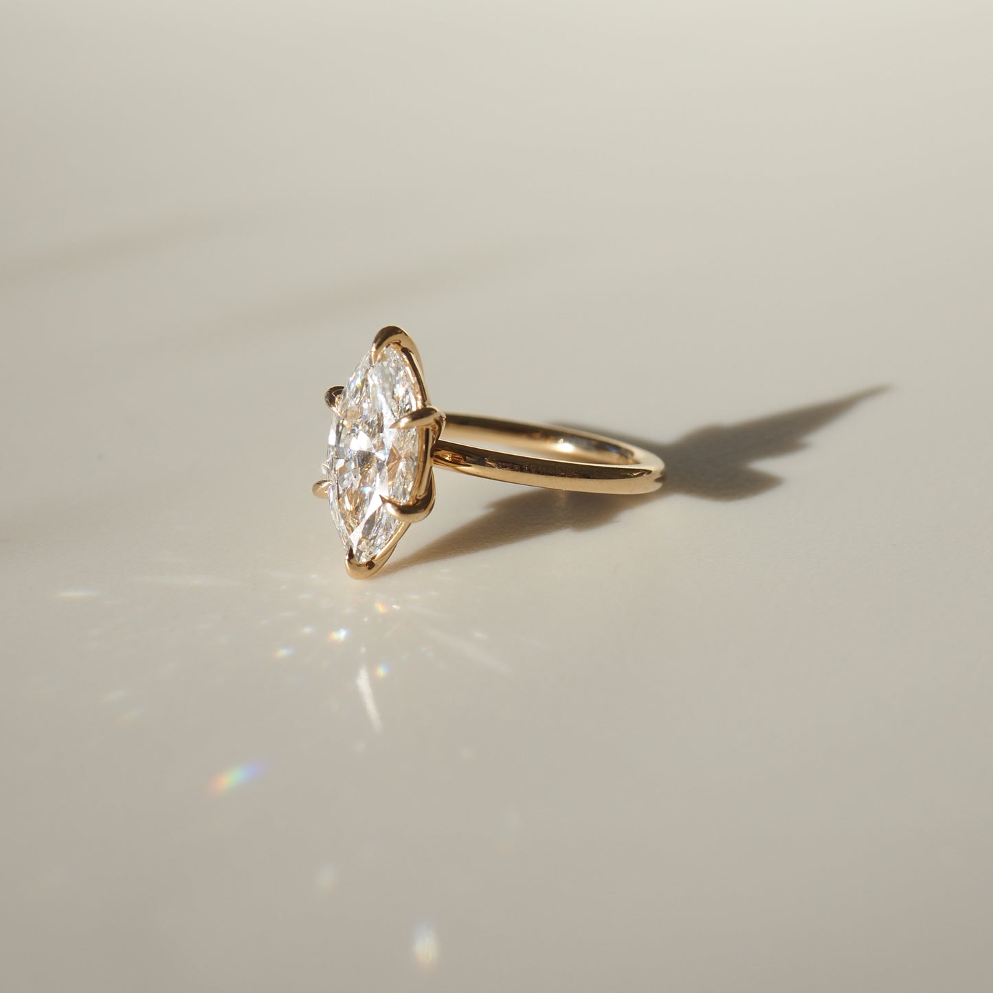 Gold ring with a marquise diamond, casting a shadow on a beige surface. The diamond sparkles in soft light, evoking luxury and elegance.