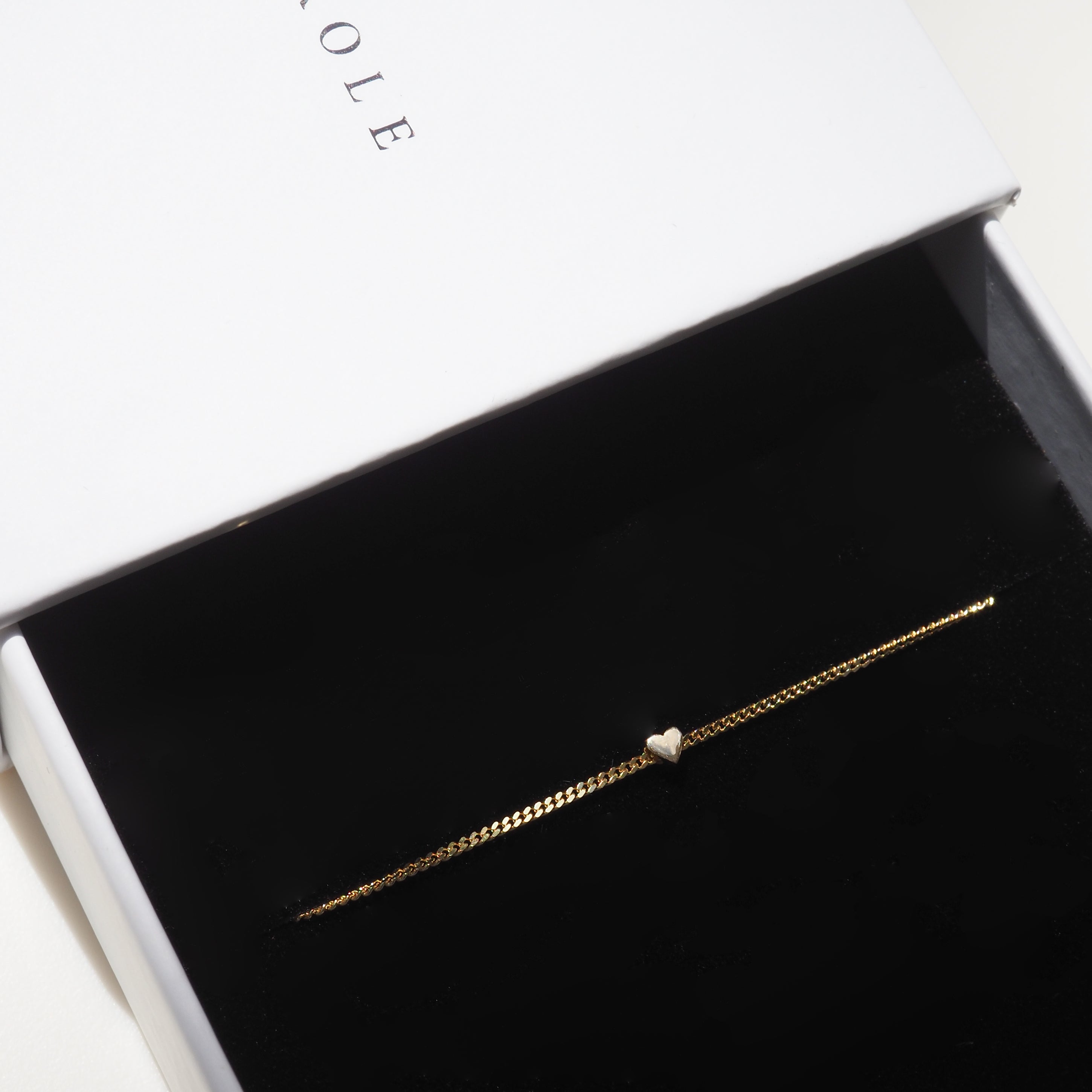 Gold bracelet with a heart charm in a black velvet box, partially open white box lid above. Luxury jewellery packaging.