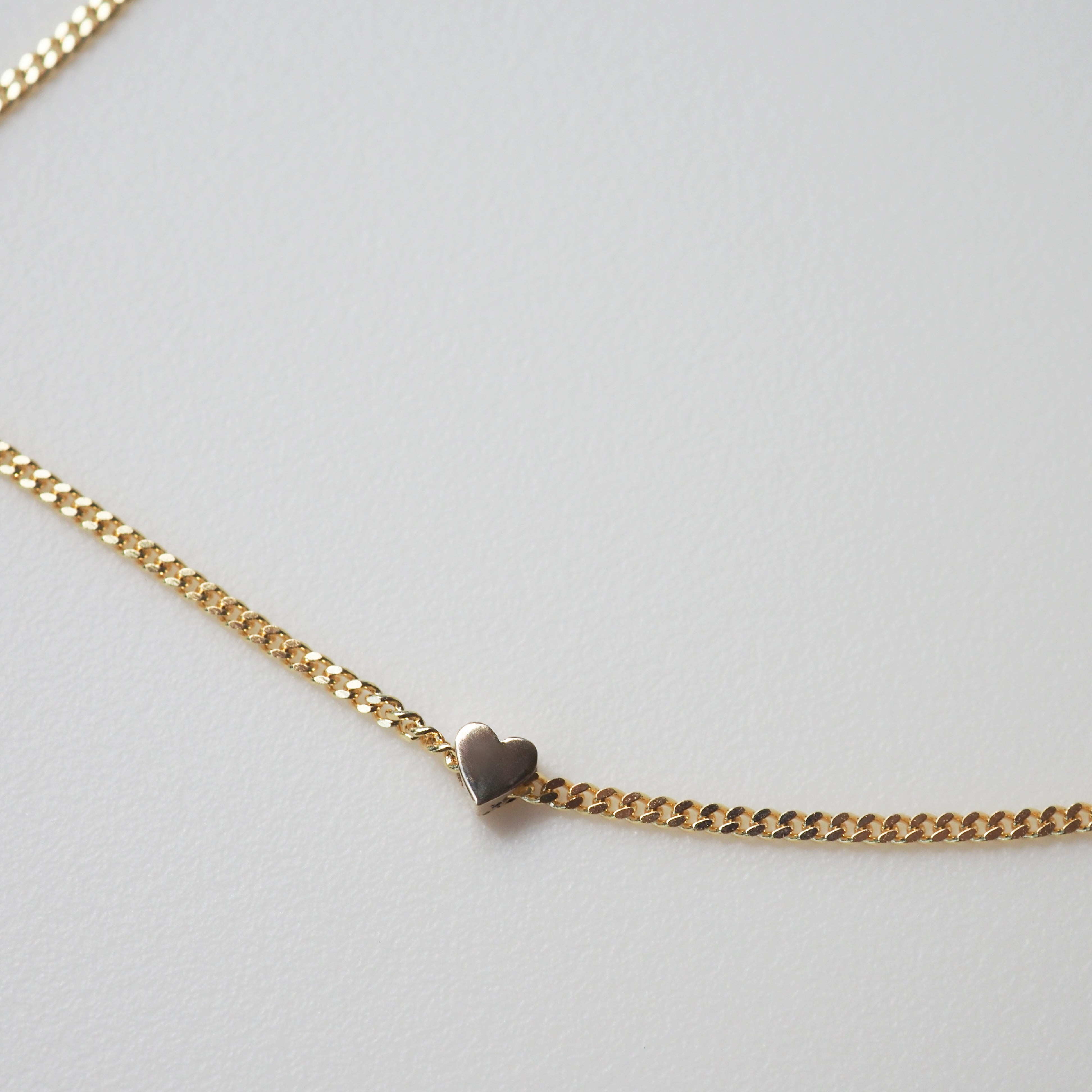 Gold chain bracelet with a small heart charm on a light grey background. Ideal for luxury jewellery lovers.