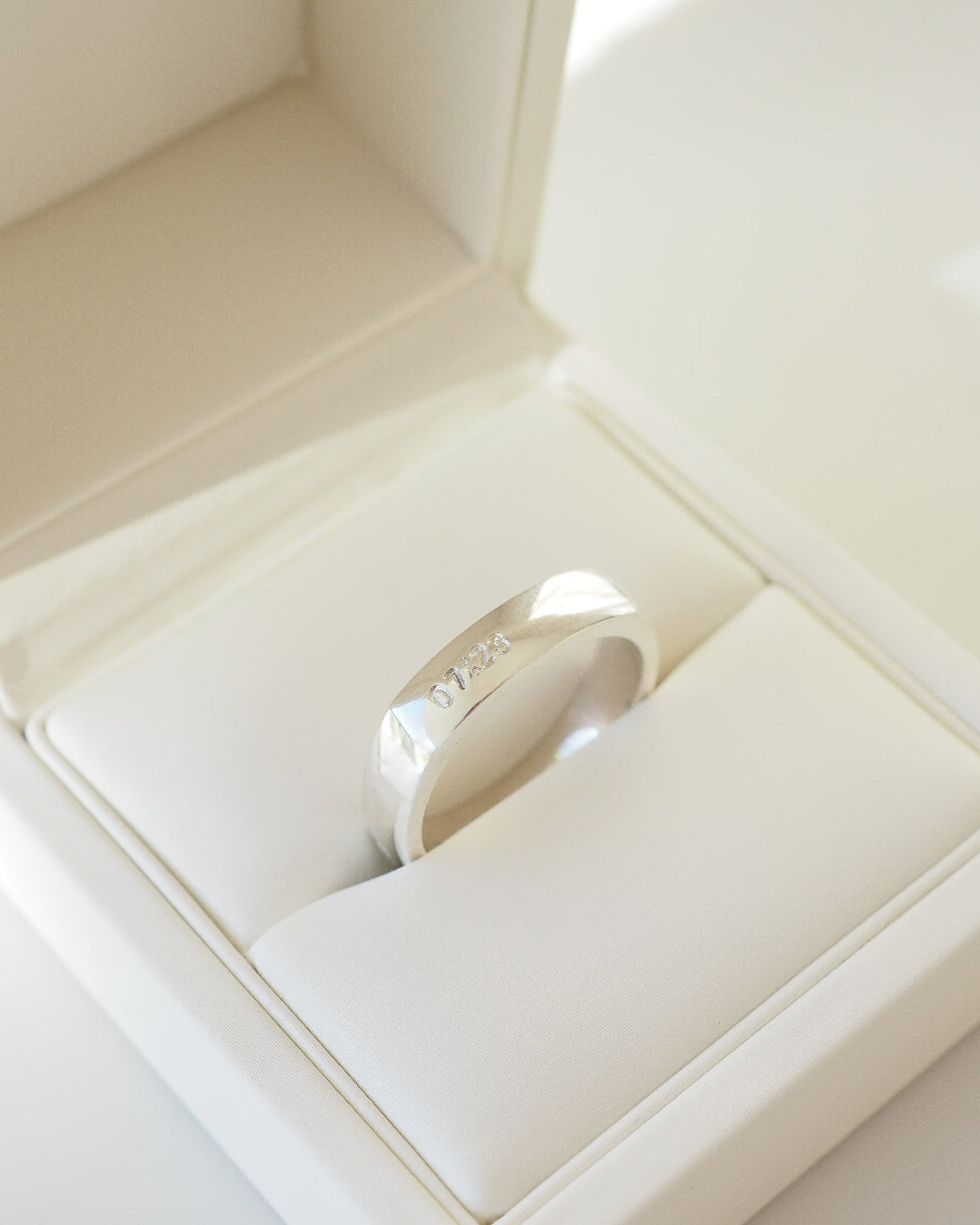 A polished silver band ring with engraved lettering, displayed inside a cream-colored jewelry box.