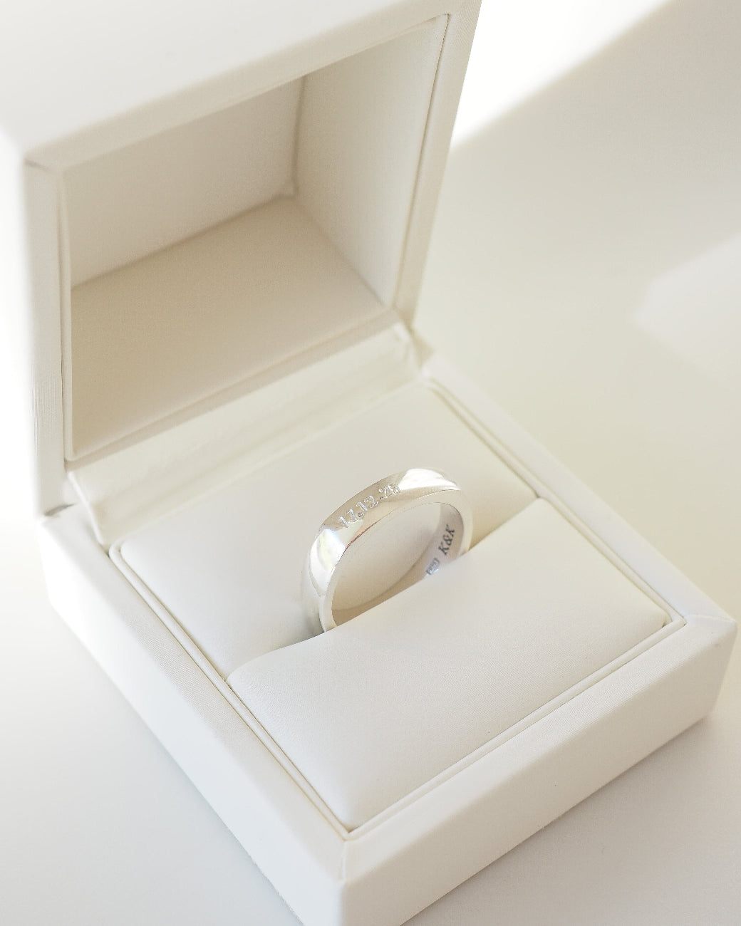A polished silver band ring displayed inside an open white jewelry box with a cream-colored velvet cushion.