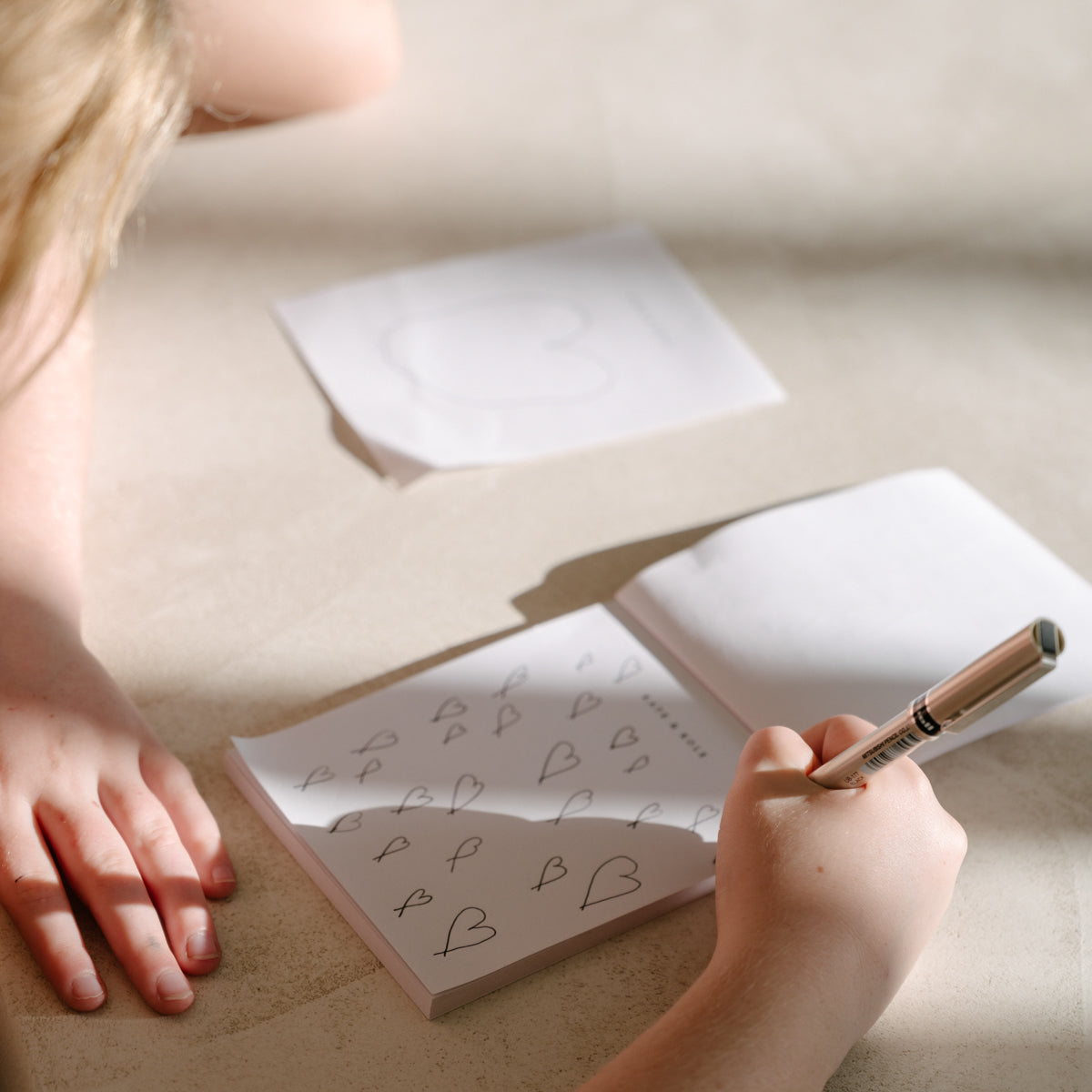 A person doodling hearts on a notepad with a pen, paper sheets scattered on a light surface. Bright, sunny setting suggests creativity.