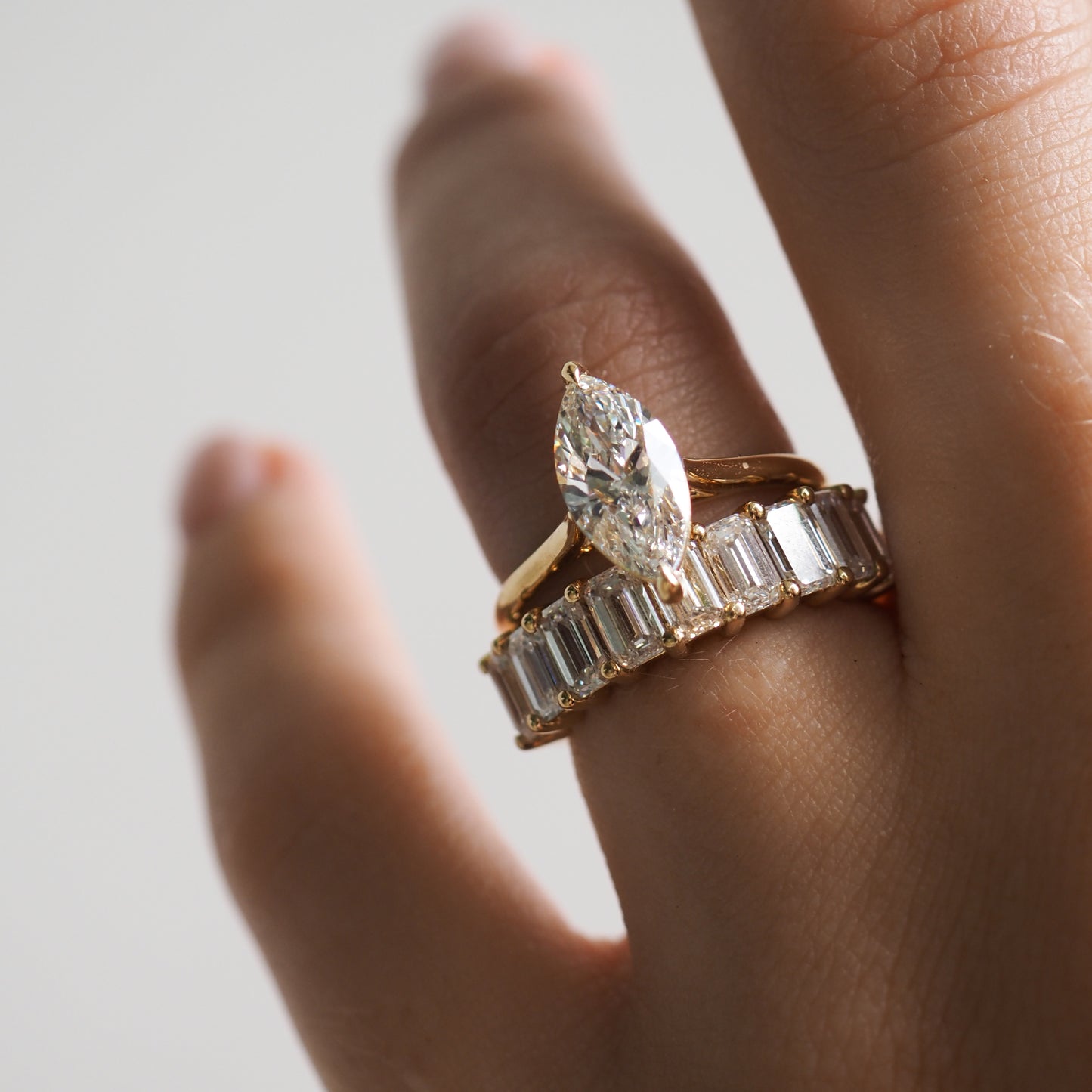 Close-up of a hand wearing a marquise-cut diamond ring atop a gold band with emerald-cut diamonds, symbolising luxury and elegance.[text overlay: 1ct Mia (top) and Emerald Lace][text colour: #000000]
