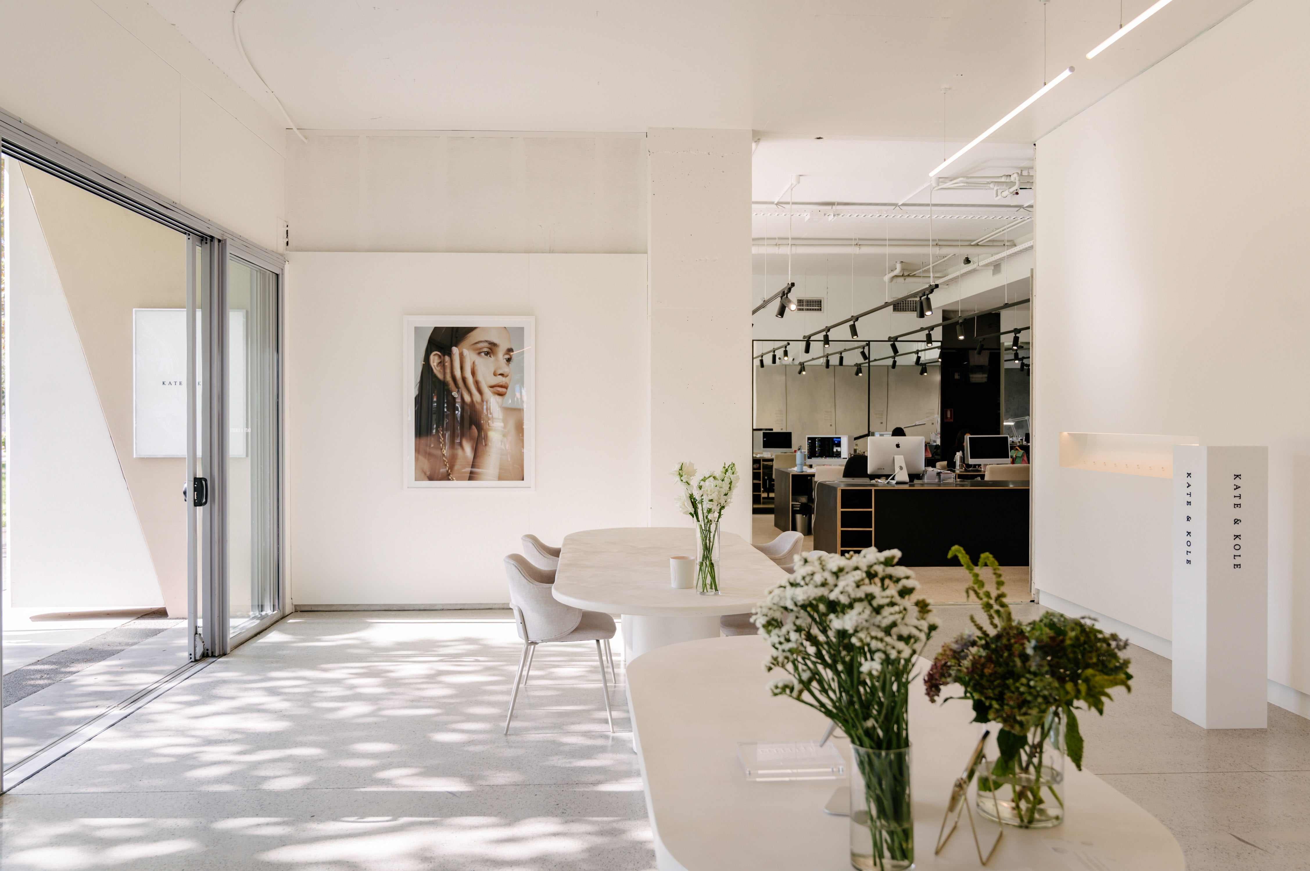 Modern jewellery showroom with minimalist decor, large photo of woman wearing ring, elegant tables, and floral arrangements. Desks visible in the background.
