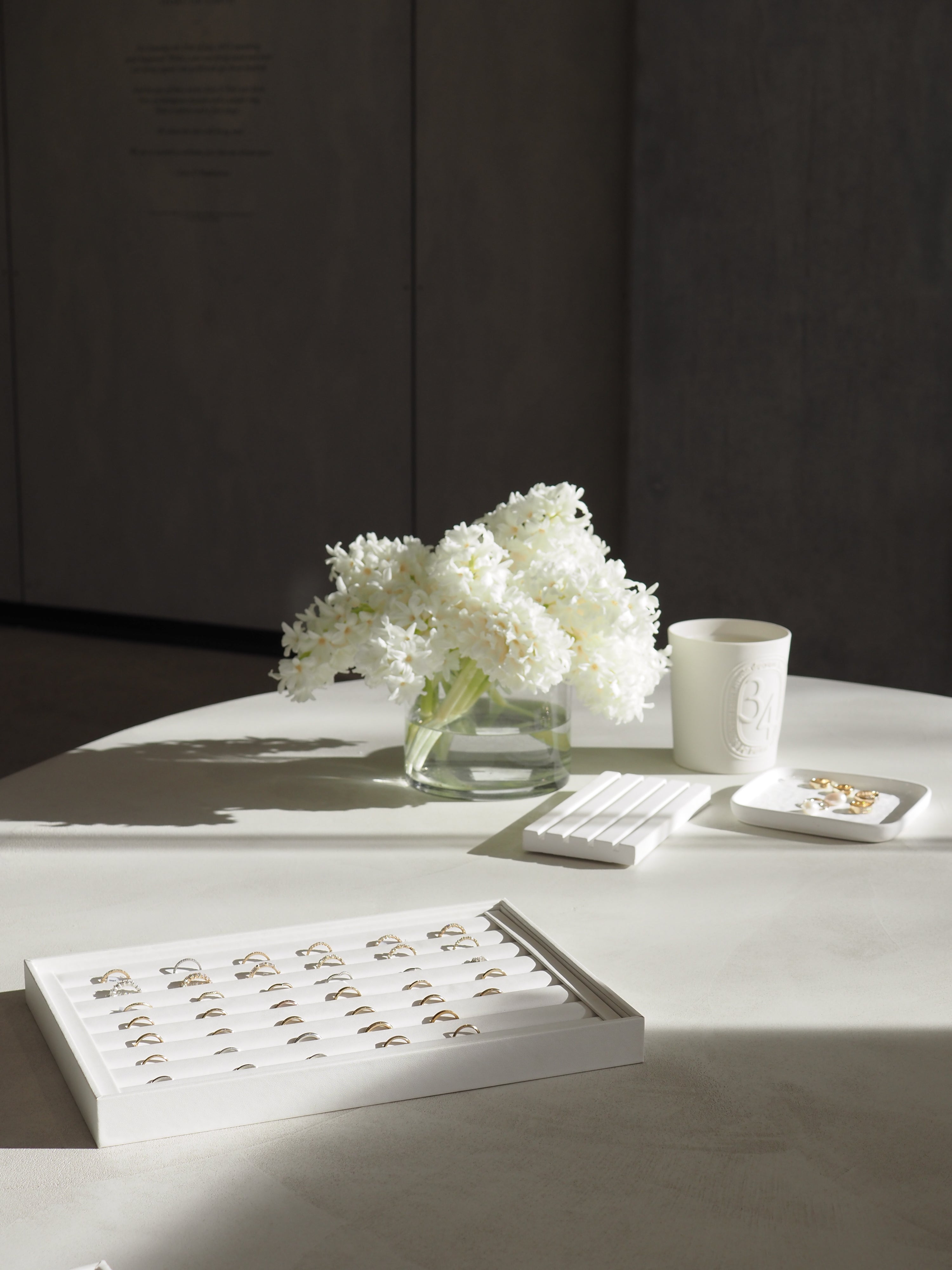 Luxurious jewellery display with gold rings on a white tray, a bouquet of white flowers, and a candle on a sunlit table.