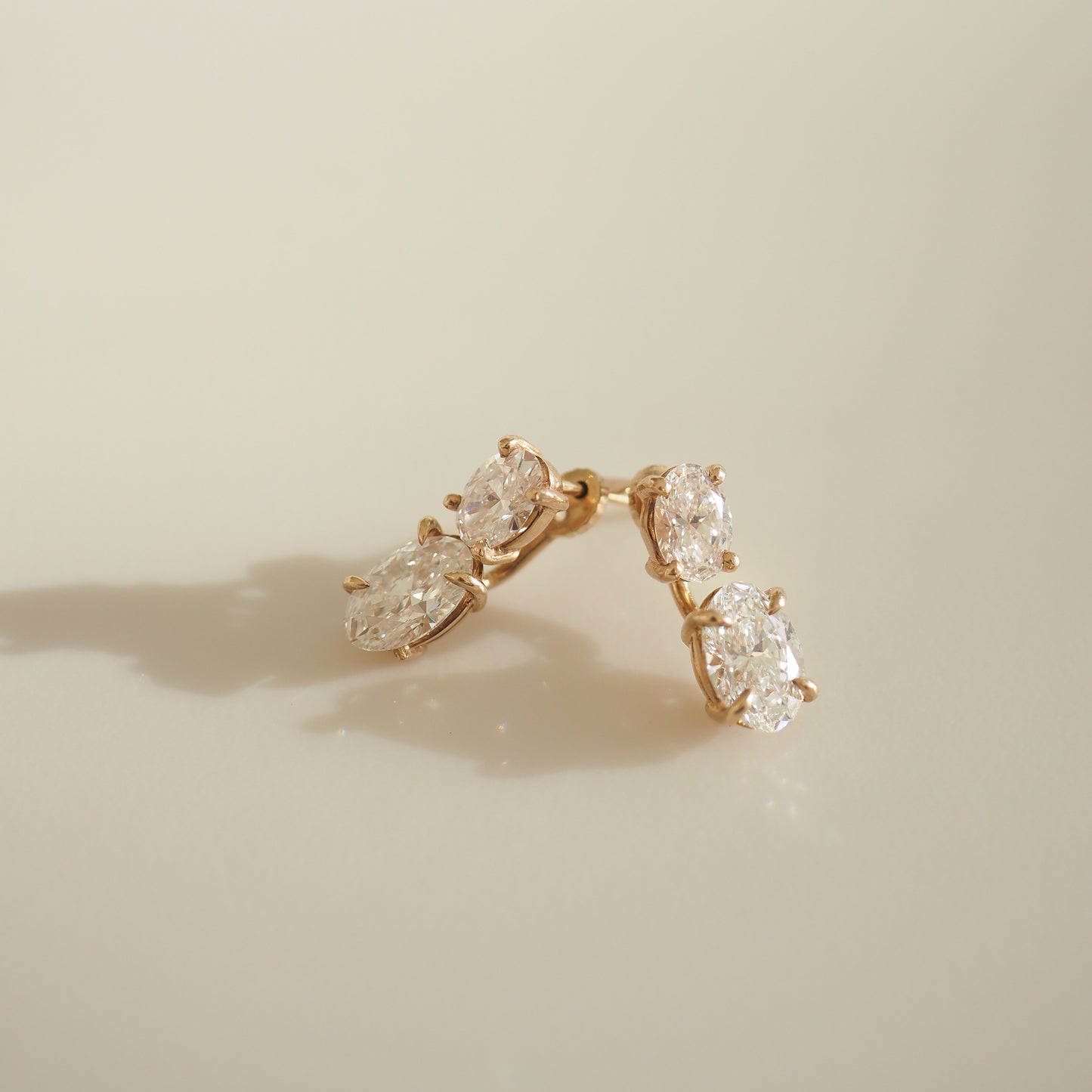 Elegant gold earrings with three sparkling oval diamonds, casting a soft shadow on a cream background. Luxurious jewellery aesthetic.