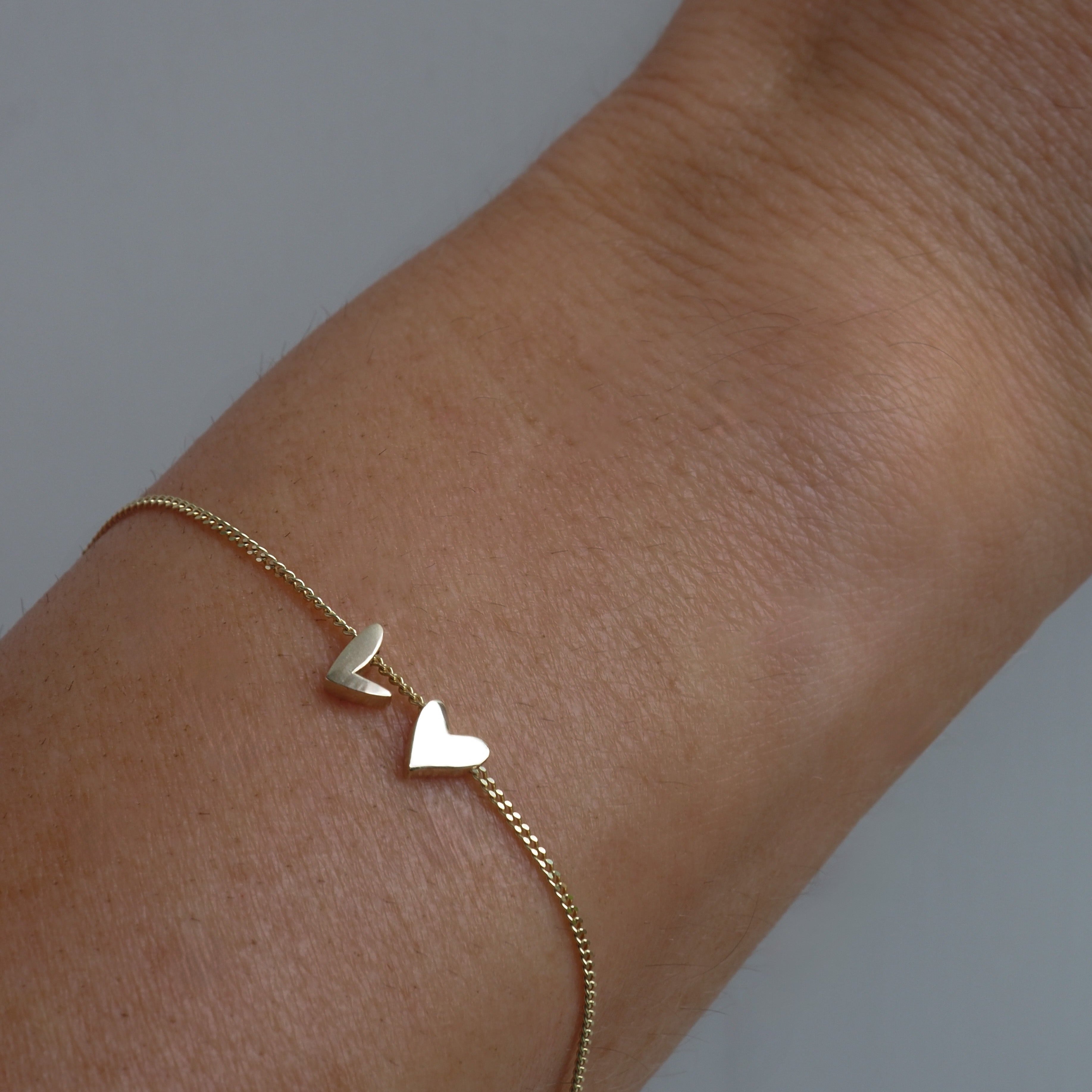 Gold heart bracelet on wrist, featuring two small heart charms on a fine gold chain. Elegant and minimalist luxury jewellery piece.[text overlay: With two charms ] 