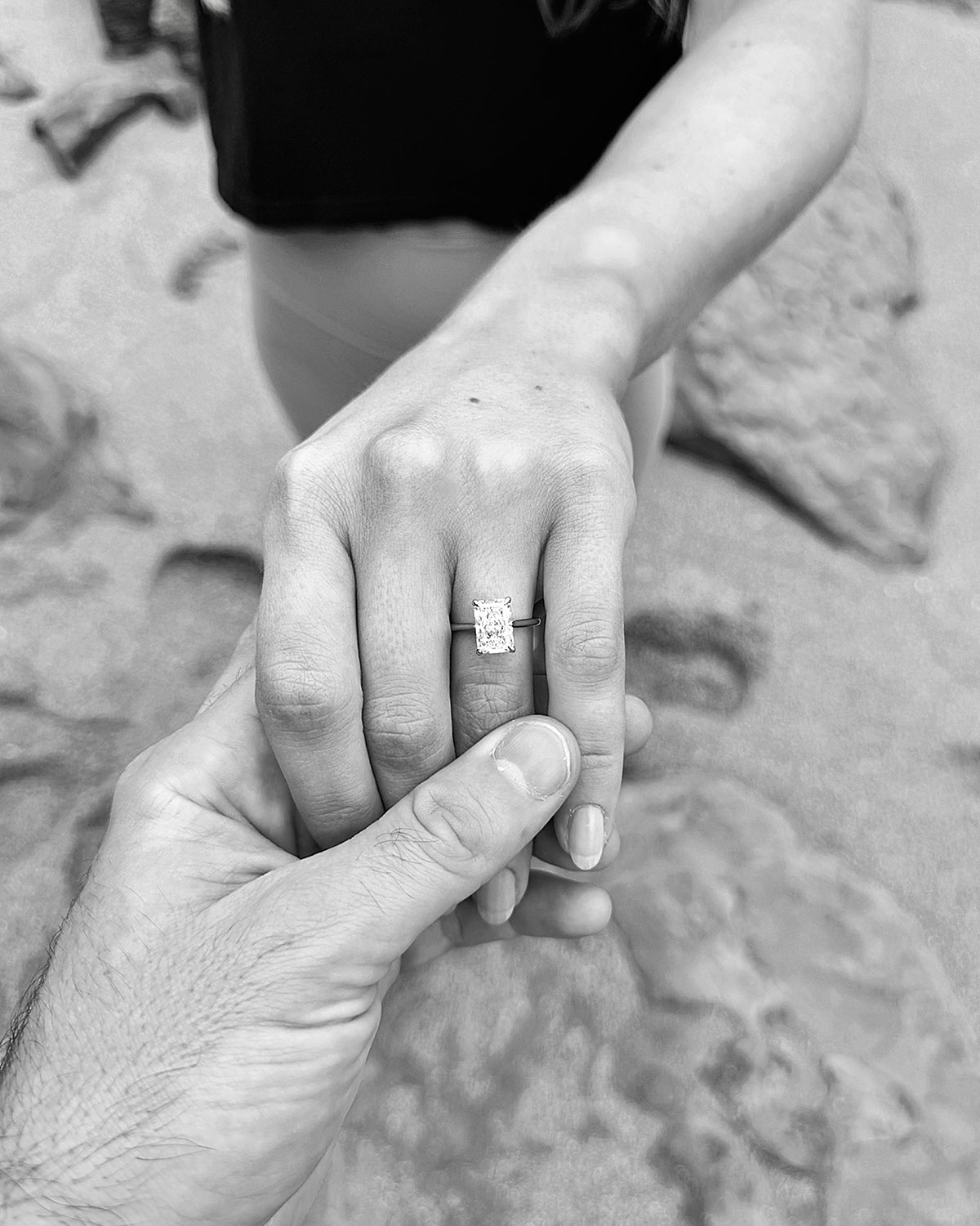 Close-up of a couple holding hands, featuring a rectangular-cut diamond engagement ring. Background shows sandy beach and rocks. Black and white.