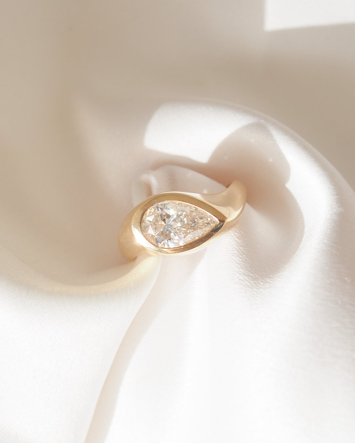 A gold ring featuring a pear-cut clear gemstone in a bezel setting, displayed on a soft white satin surface.