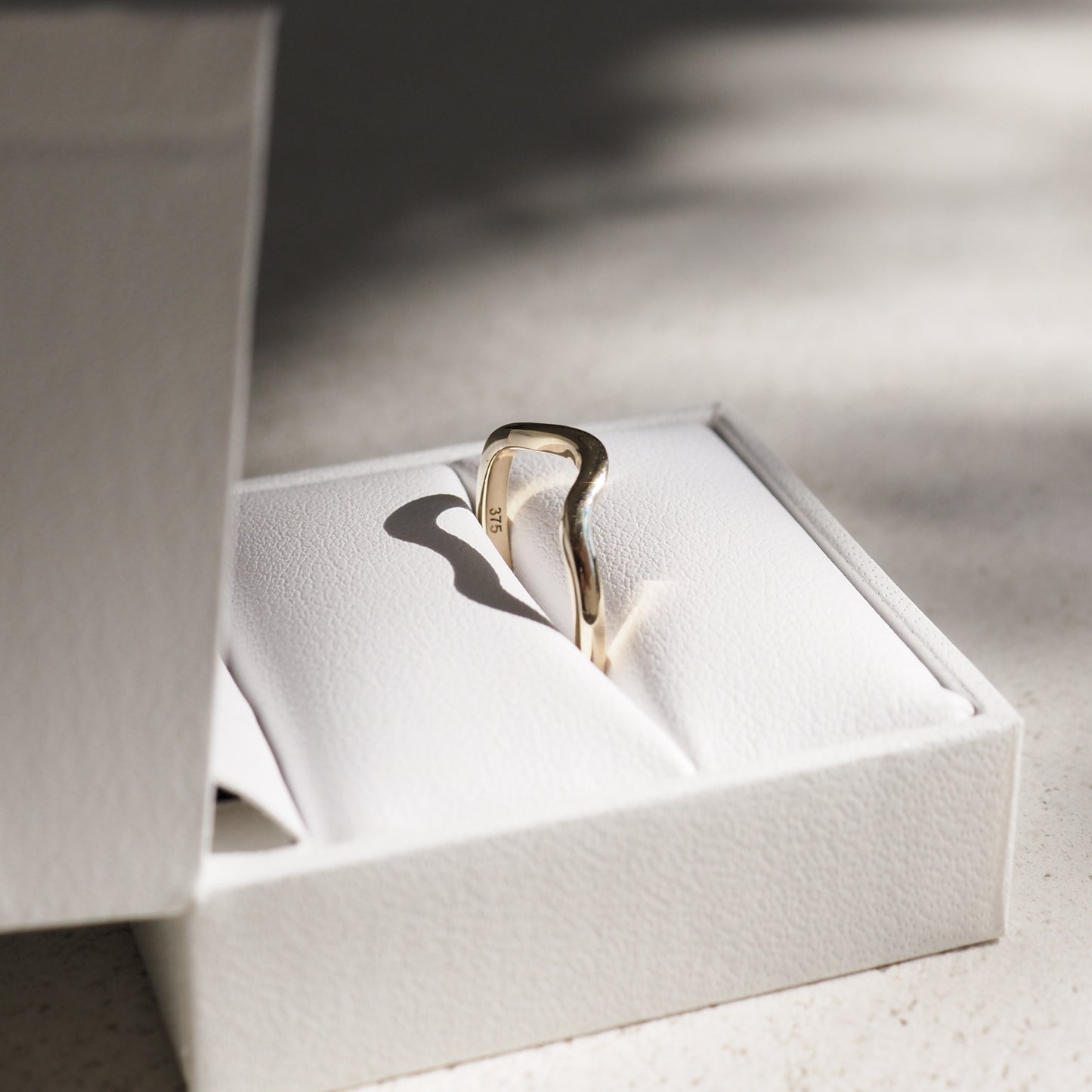 Elegant gold ring with a wavy design resting in a white jewellery box, casting a soft shadow. Natural light enhances its luxurious appeal.
