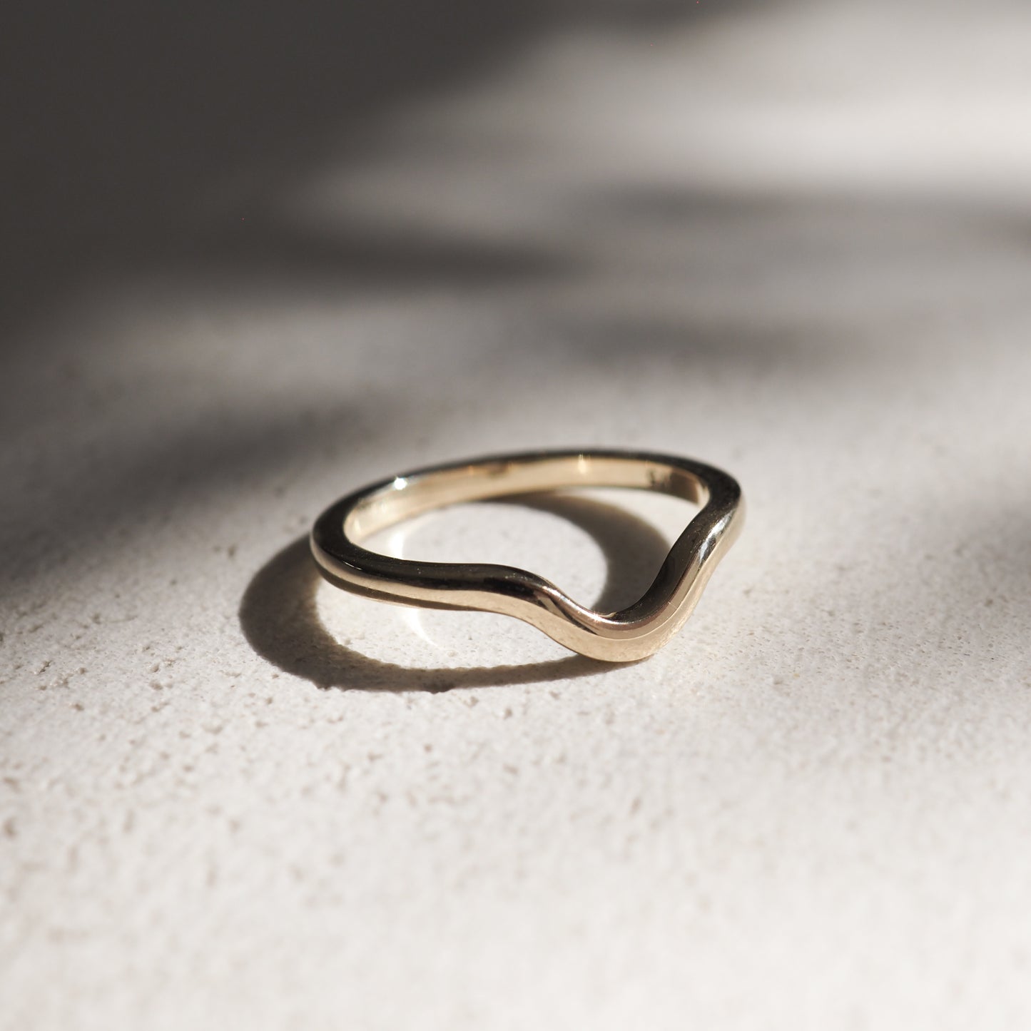 Gold wave ring on a textured surface, bathed in soft, natural light. Elegant curves reflect minimalist, luxury jewellery design.