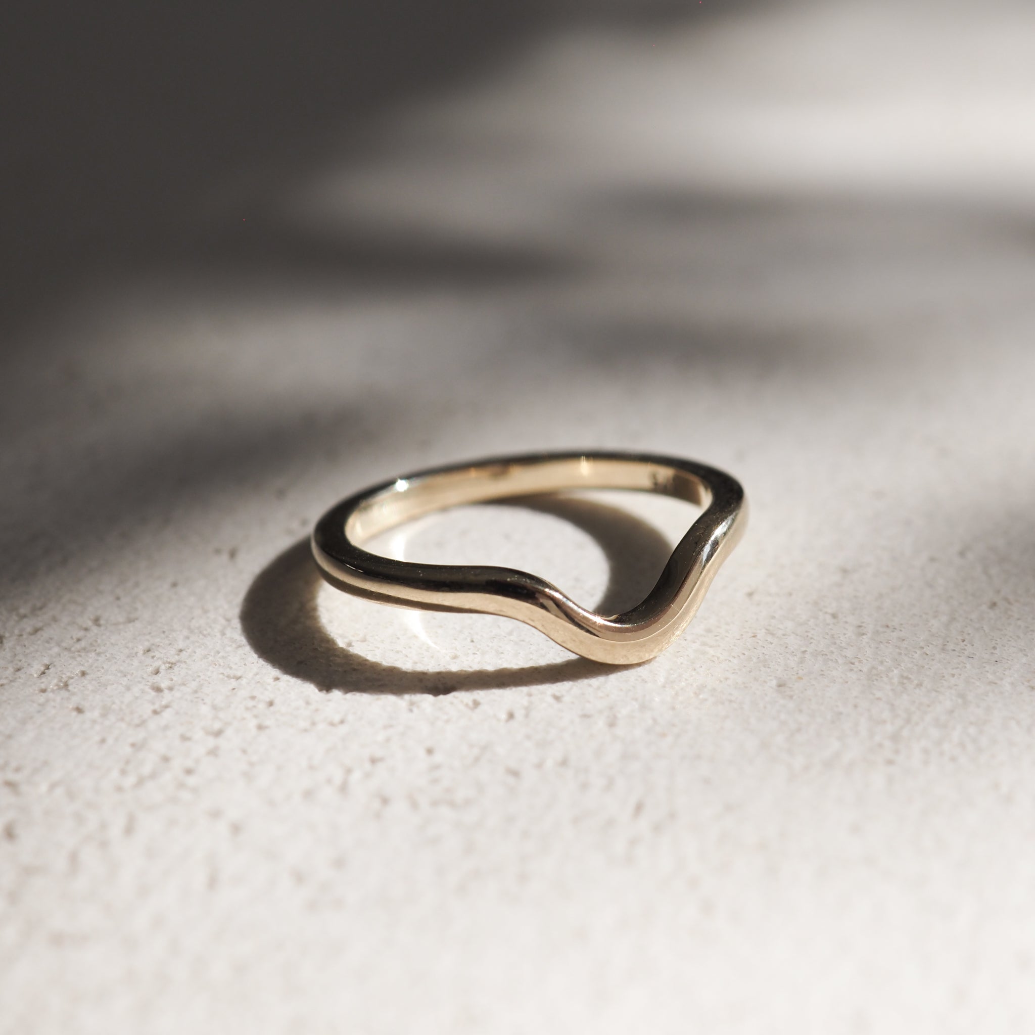 Gold wave ring on a textured surface, bathed in soft, natural light. Elegant curves reflect minimalist, luxury jewellery design.
