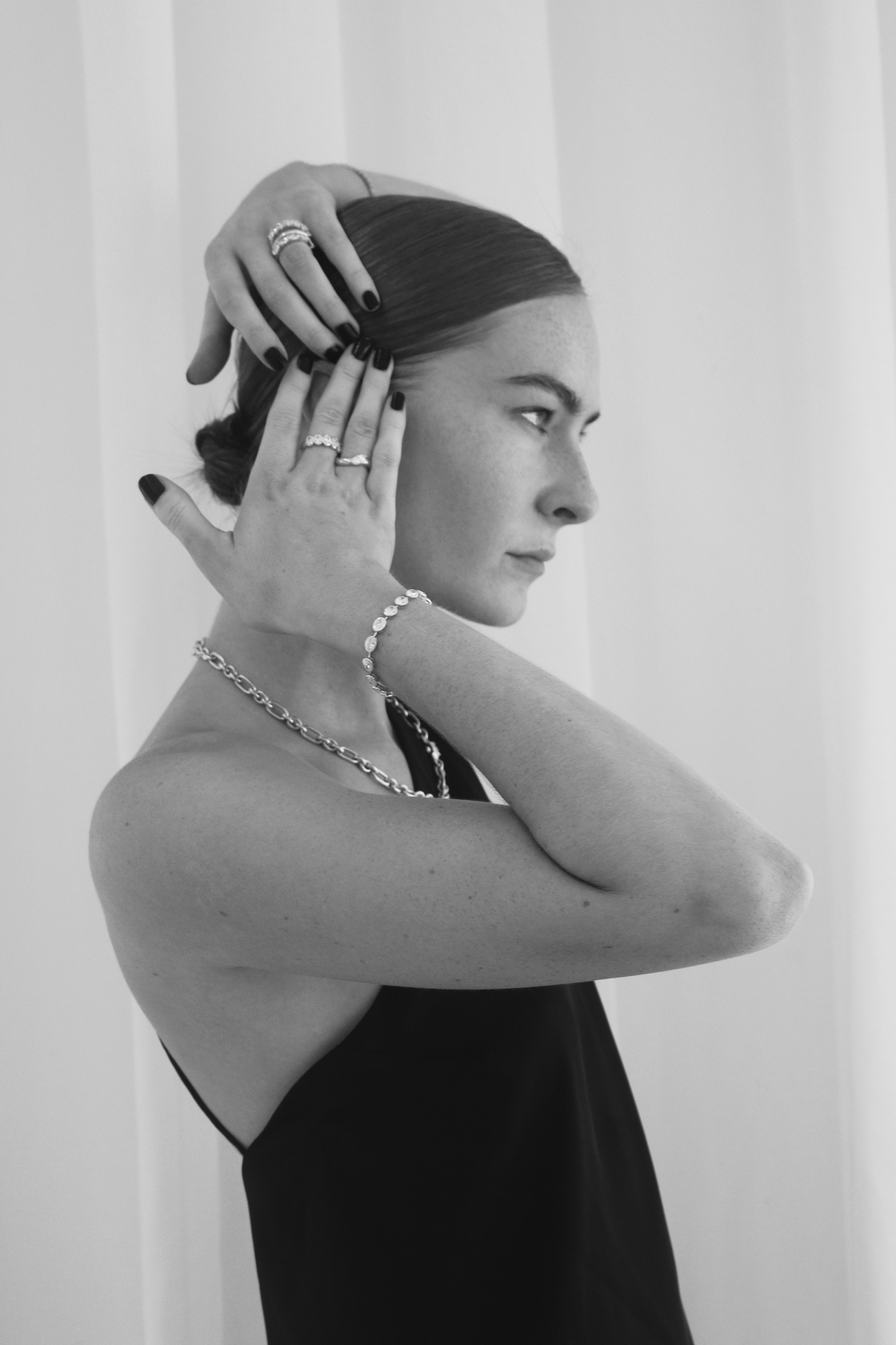 Woman in elegant black dress showcases luxury jewelry: diamond rings, bracelet, and necklace, against a soft curtain backdrop.