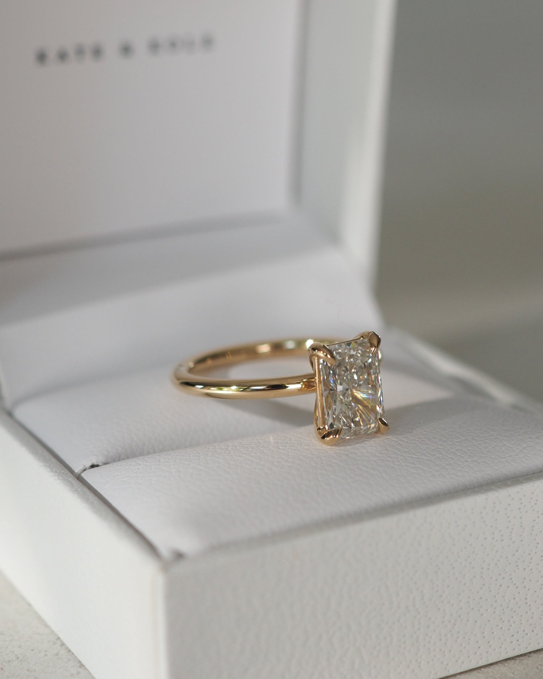 Gold engagement ring with a radiant-cut diamond in a white jewellery box. Elegant and luxurious design, perfect for proposals.