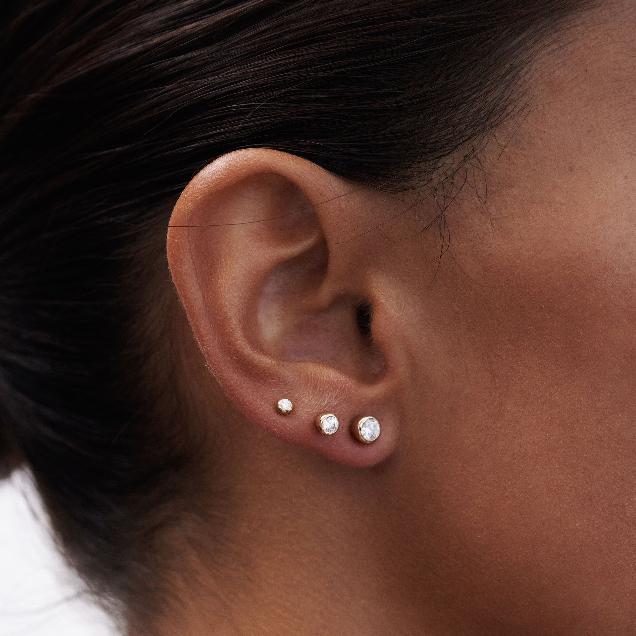 Close-up of ear featuring three diamond studs in varying sizes on the lobe, showcasing elegant luxury jewellery styling.
[text overlay: Left to right: Small, Medium and Large]