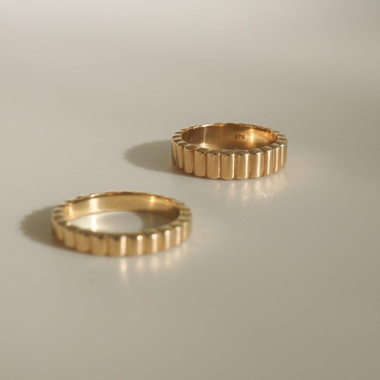 Two ribbed gold rings on a light surface, one standing upright showing "375" hallmark inside. Luxurious jewellery, minimalistic design.