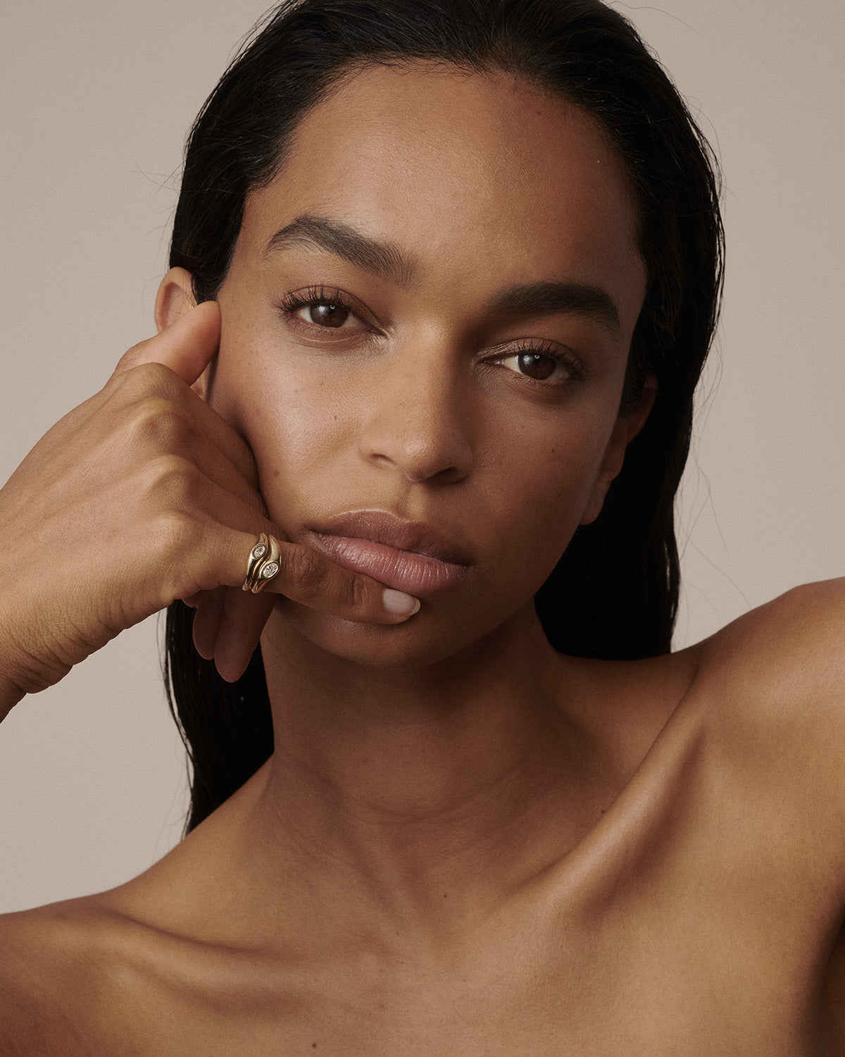 A close-up portrait of a woman with dark hair and warm skin tones, wearing a gold ring on her finger, set against a solid beige background.