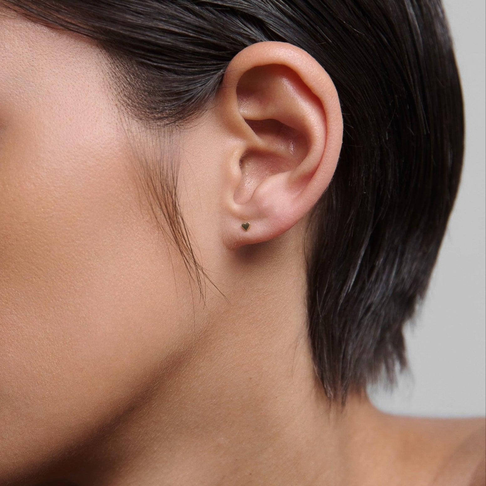 Profile of a person with slicked-back hair wearing a subtle, small stud earring. Skin appears smooth and makeup is minimal.