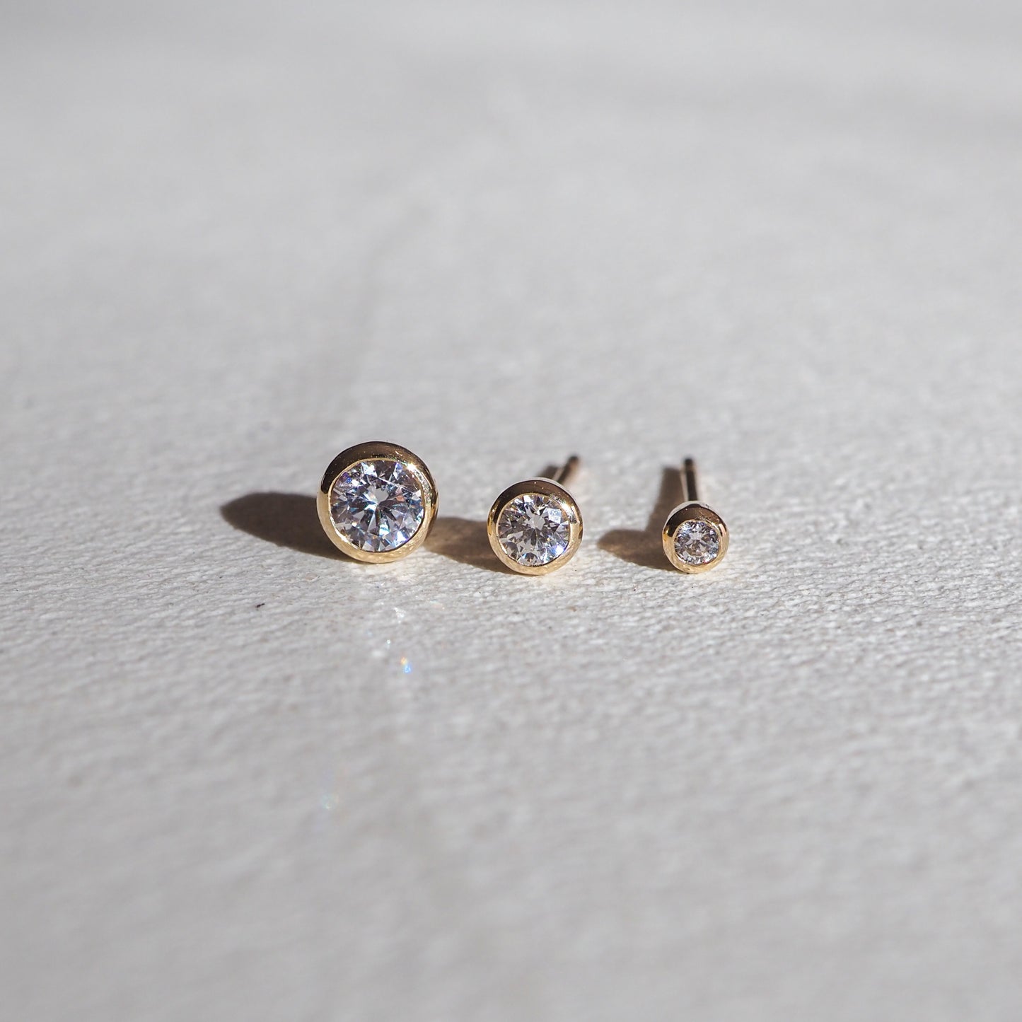 Three gold bezel-set diamond studs arranged by size on a textured white surface, showcasing luxury jewellery craftsmanship.