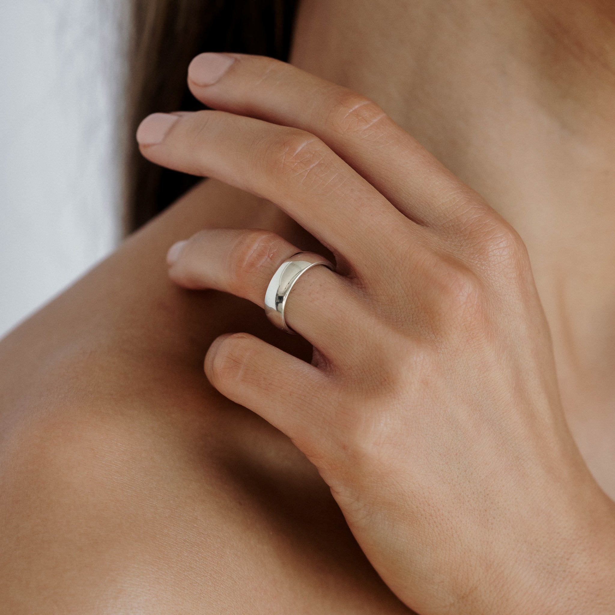 Close-up of a hand with nude nails gently touching a bare shoulder, showcasing a sleek, polished silver ring on the ring finger.