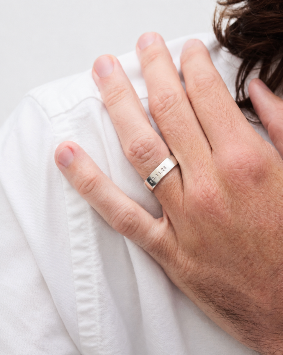 A close-up of a man's hand resting on his white shirt, wearing a silver band ring engraved.