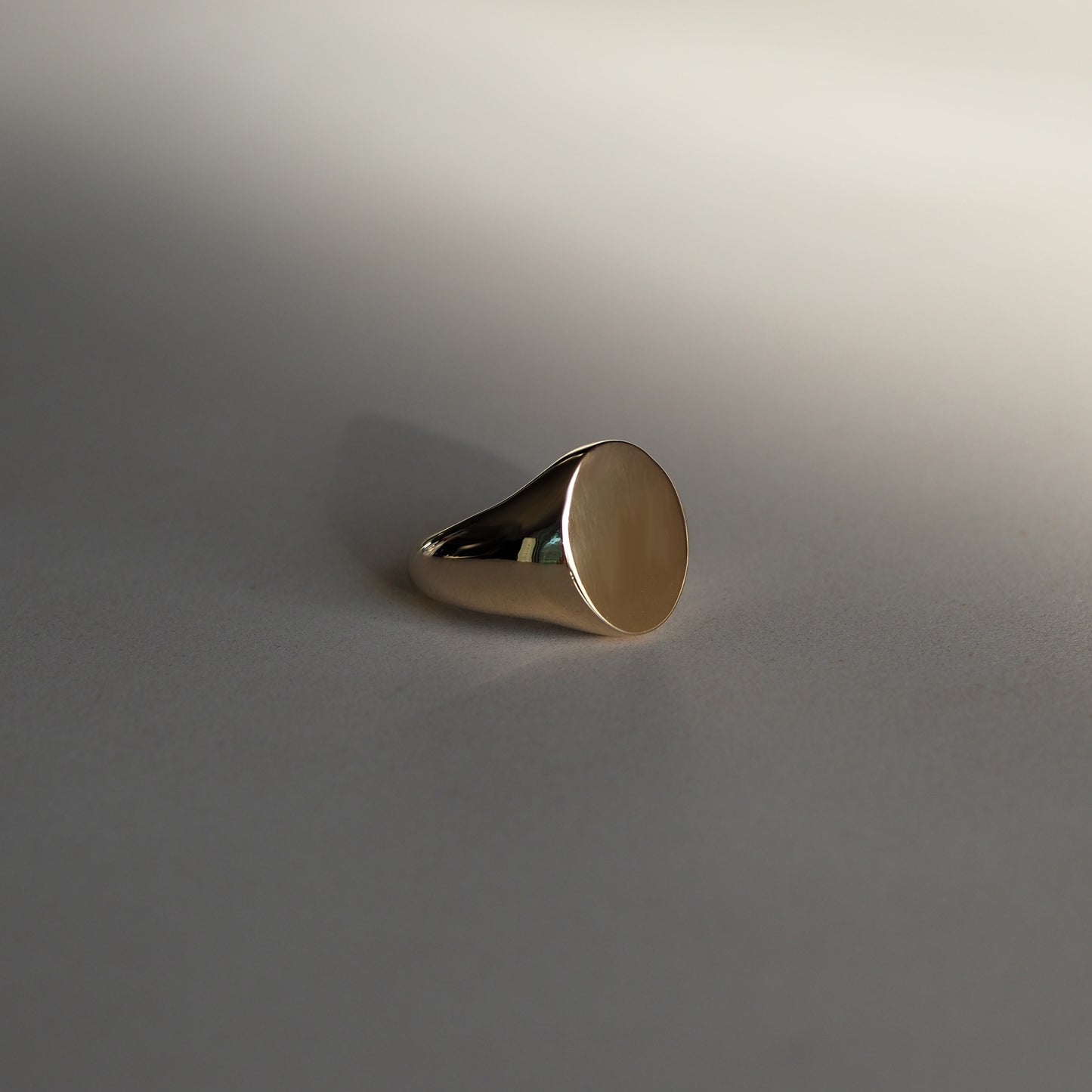 Elegant gold signet ring with a smooth, polished face on a soft grey background, showcasing high-end luxury jewellery design.