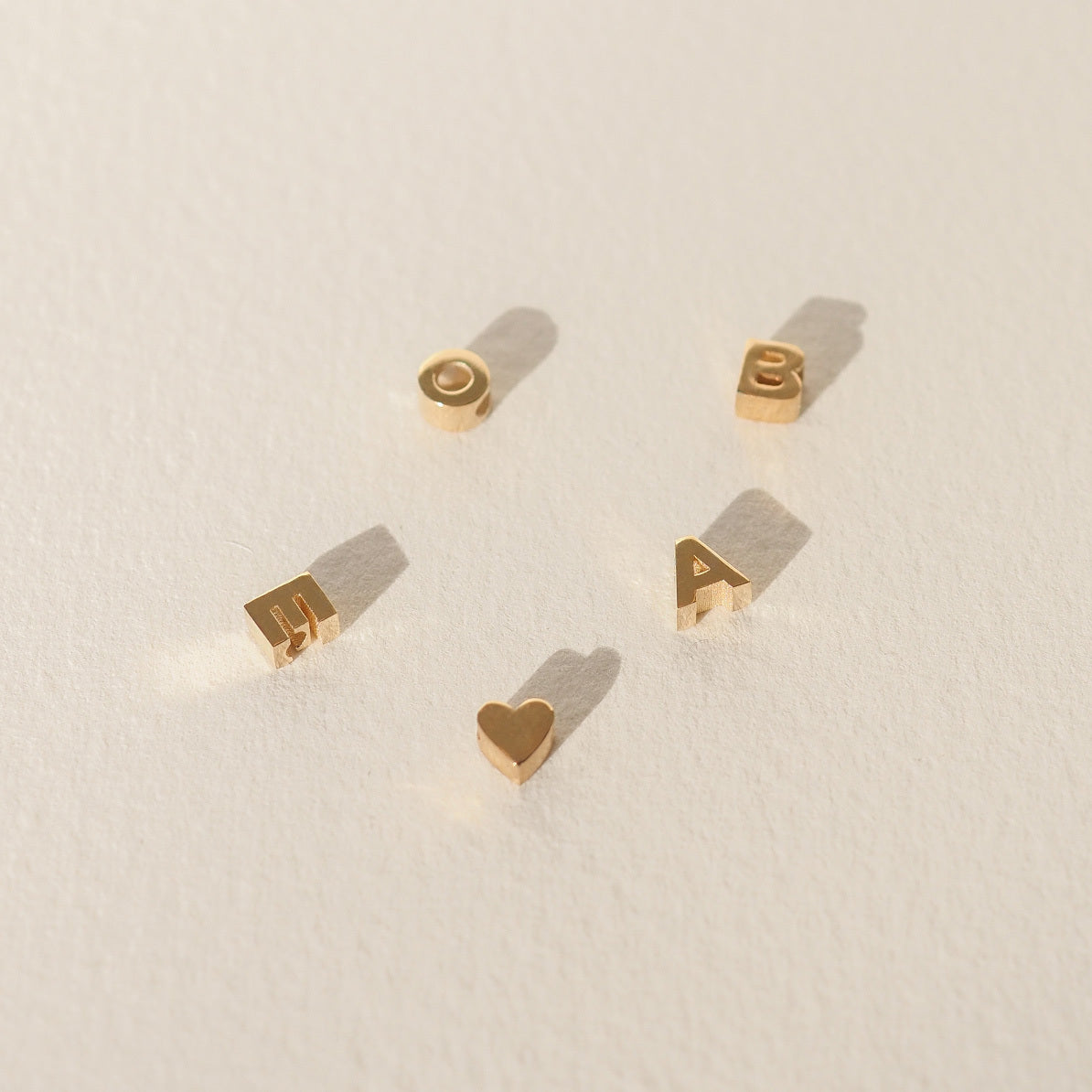 Gold alphabet and heart charms arranged on a light background, casting soft shadows. Minimalist luxury jewellery display.