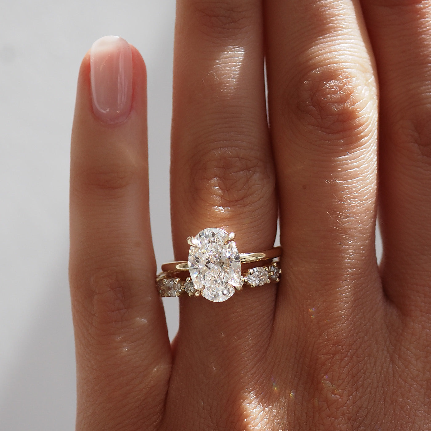 Hand with gold engagement ring featuring a large oval diamond, accompanied by a diamond-studded band, set in sparkling sunlight.