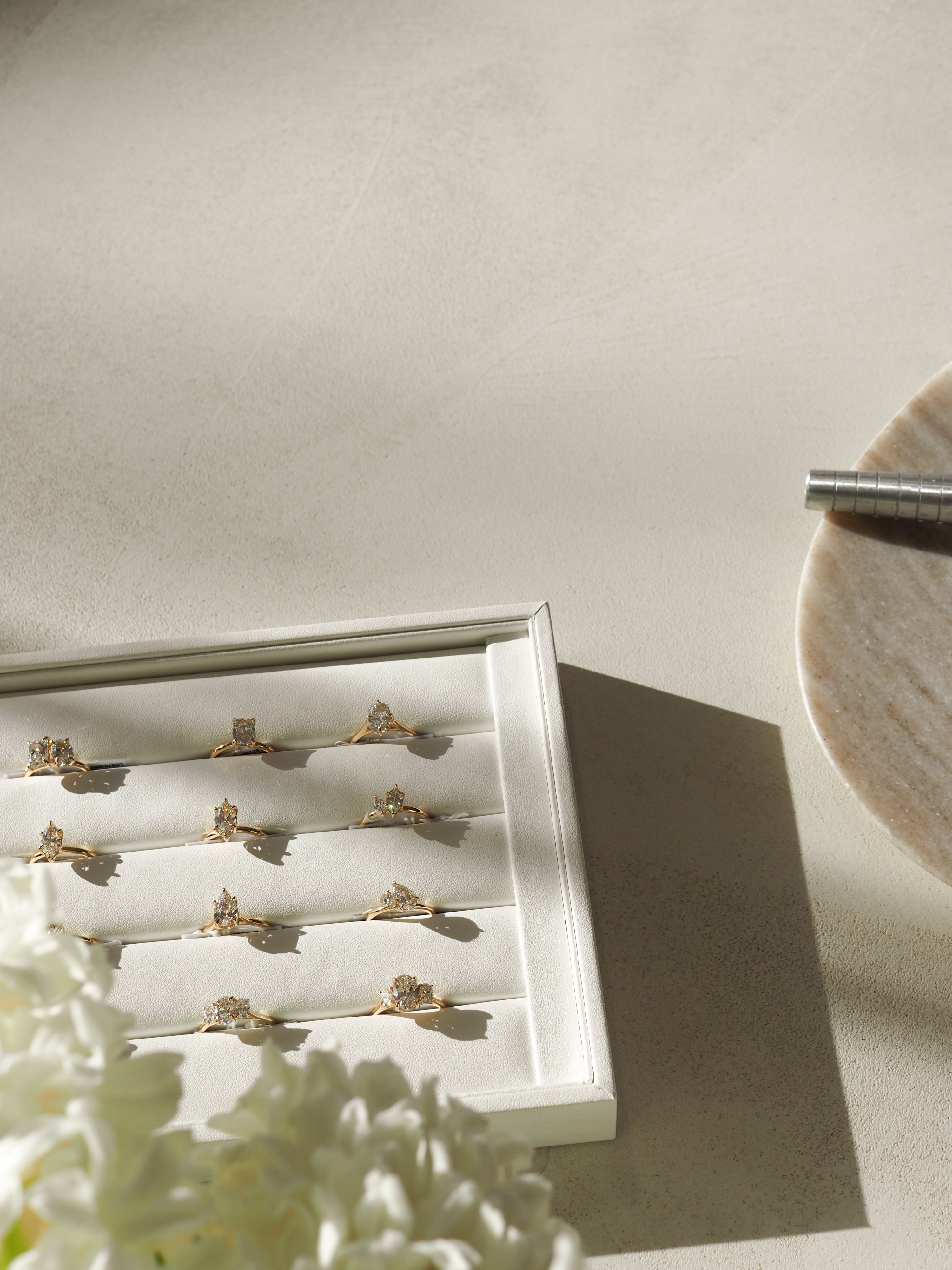 A tray of luxury diamond rings on a white surface, with a ring sizing tool and white flowers nearby. Sunlight casts soft shadows.