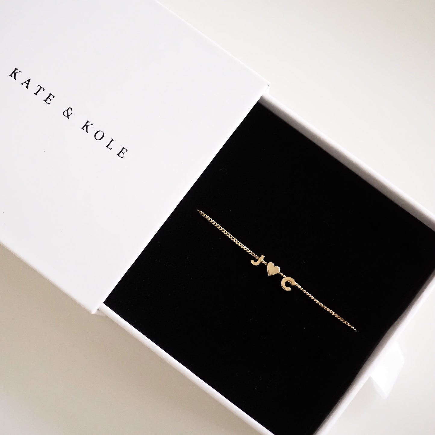 Gold bracelet with initials "J♥C" in a black velvet box, branded "KATE & KOLE." Elegant, luxury jewellery.