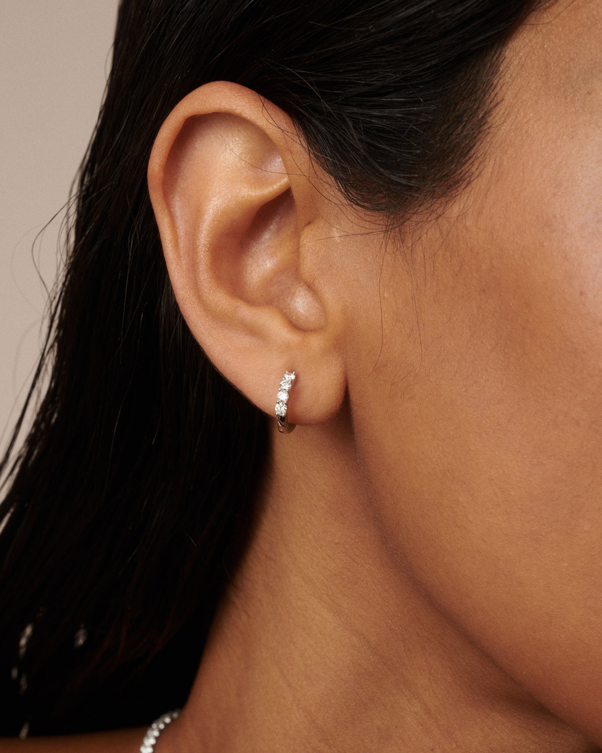 A close-up of a model's ear wearing a small silver huggie hoop earring adorned with a row of clear diamonds.