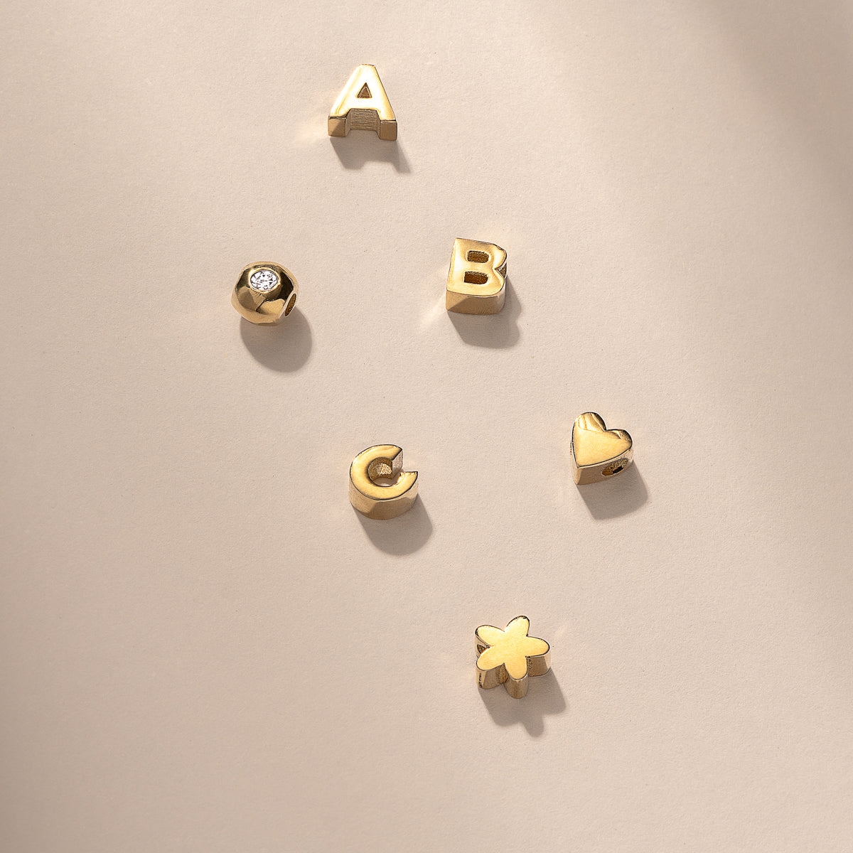 Gold letter charms A, B, C, and symbols including a gemstone O, star, and heart on a beige background; luxury jewellery accents.