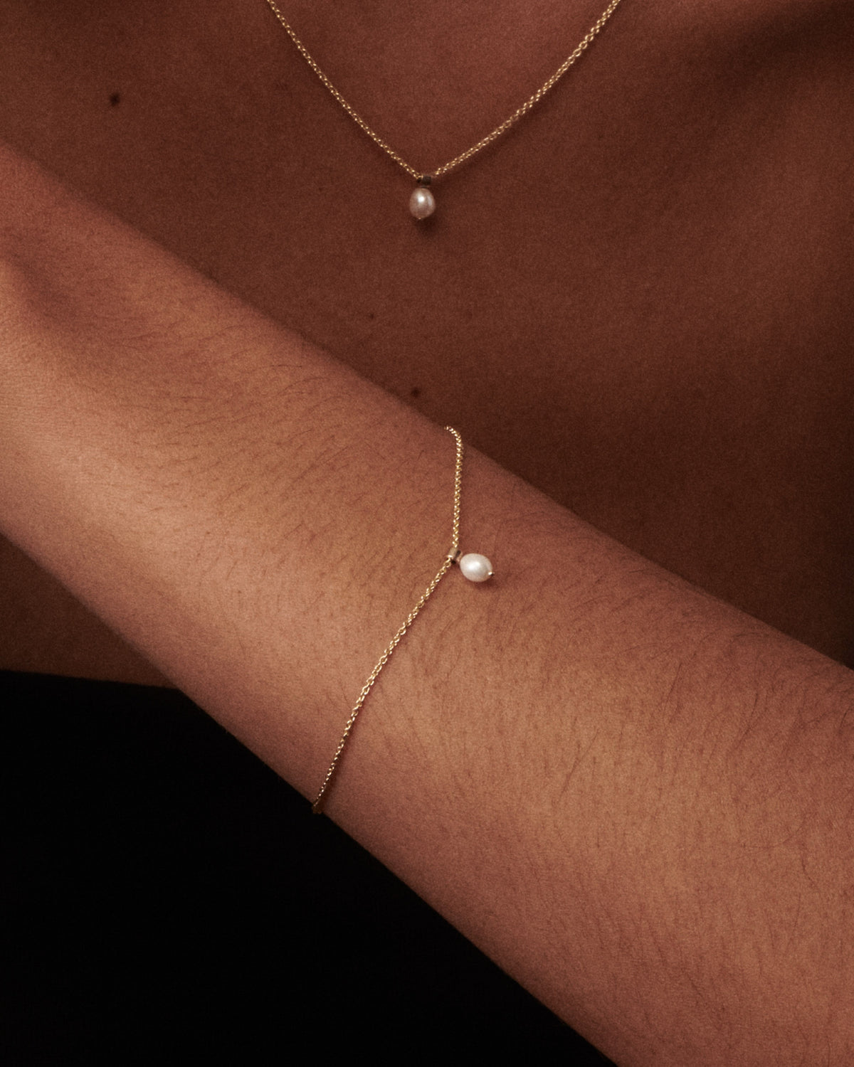 A close-up of a person's arm and chest wearing a delicate gold chain bracelet with a single white pearl, with a matching gold necklace also visible.