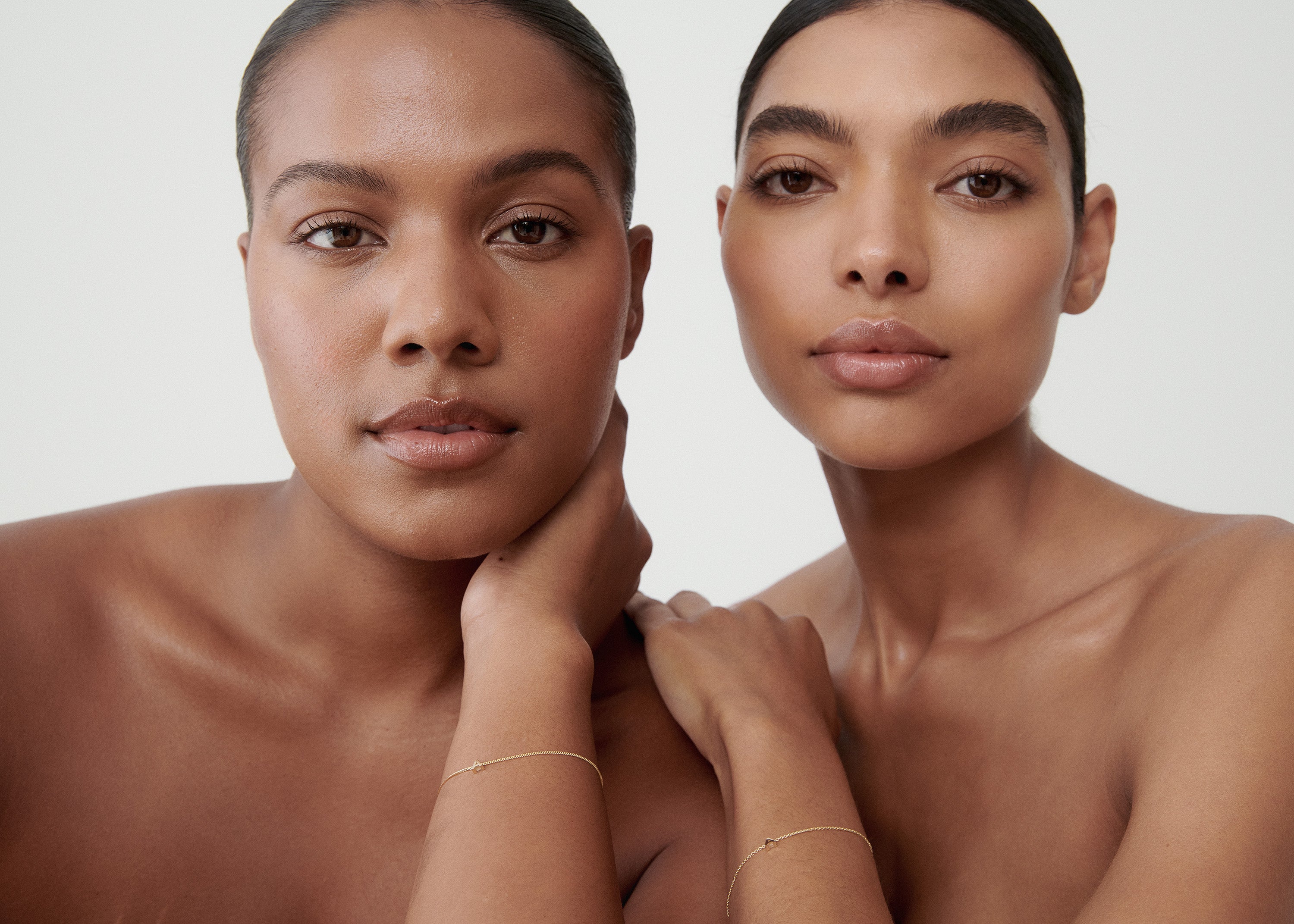 Two women with sleek hair models minimalistic gold jewellery against a neutral background, focusing on luxury and elegance.