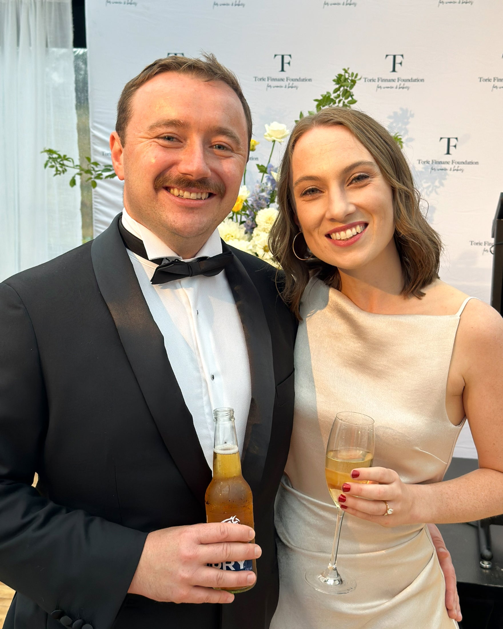 A man in a tuxedo and a woman in a satin dress smile at a formal event, holding drinks, with a floral backdrop and branded wall behind them.