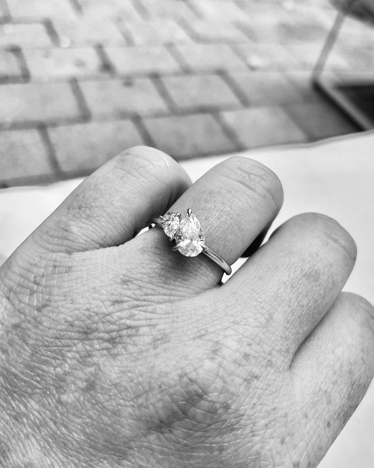 Close-up of a hand wearing a luxury three-stone diamond engagement ring on a metal band, set against a blurred, outdoor background.