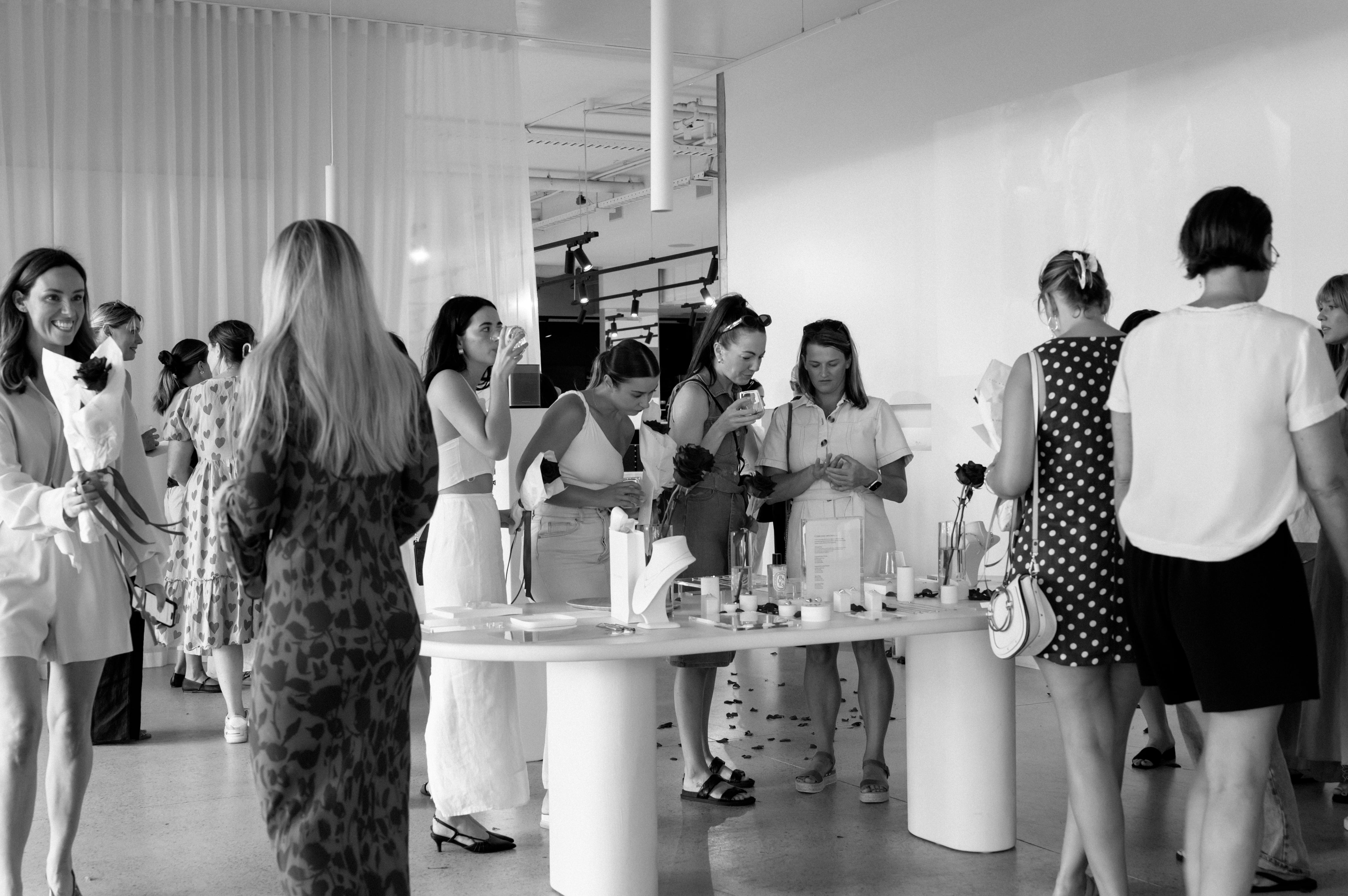 A stylish gathering in a modern jewellery showroom. Guests in chic outfits mingle around a central display table.