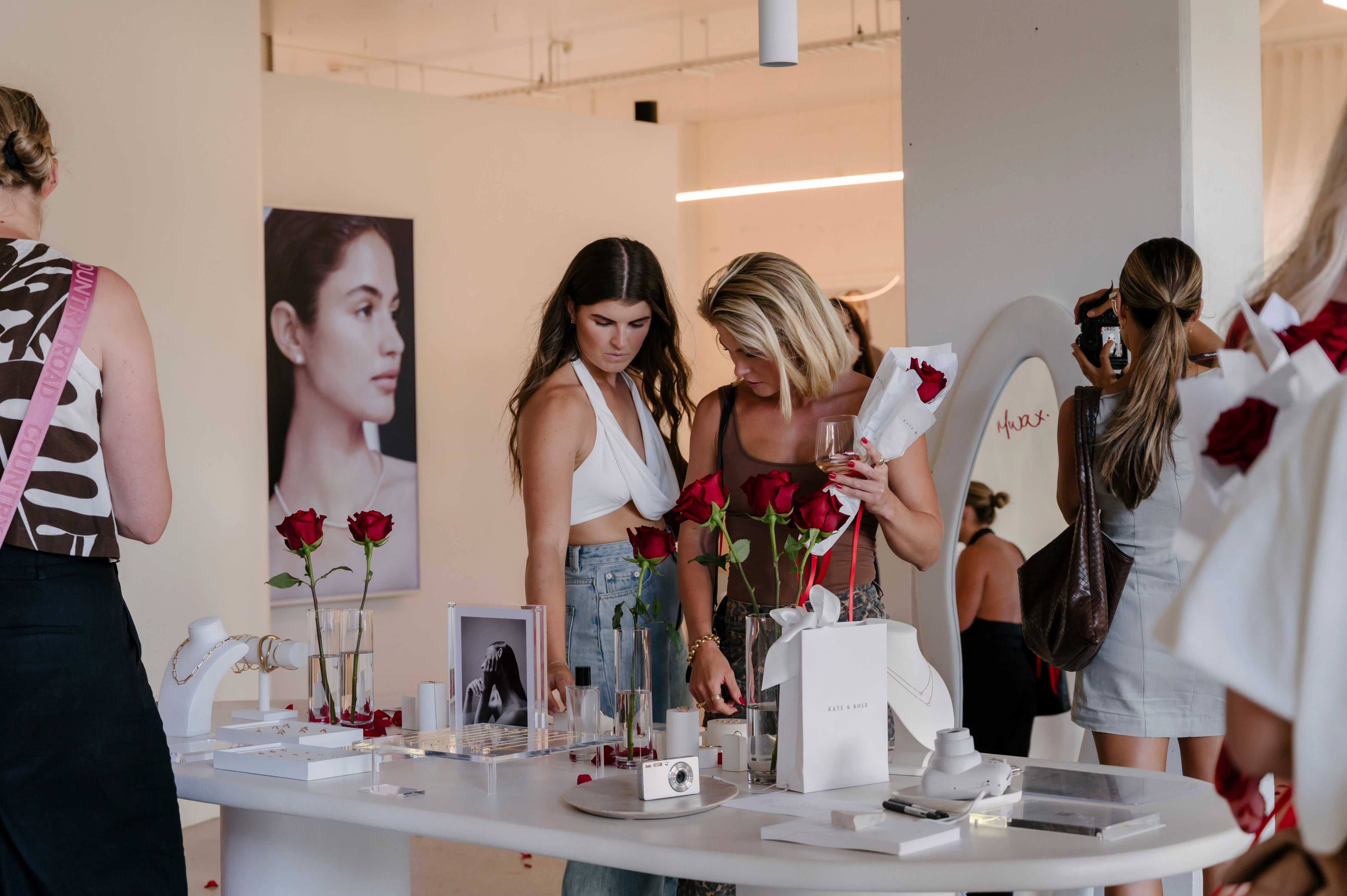 Women examine jewellery on a display table adorned with red roses in a boutique. A large portrait and elegant lighting enhance the luxurious setting.