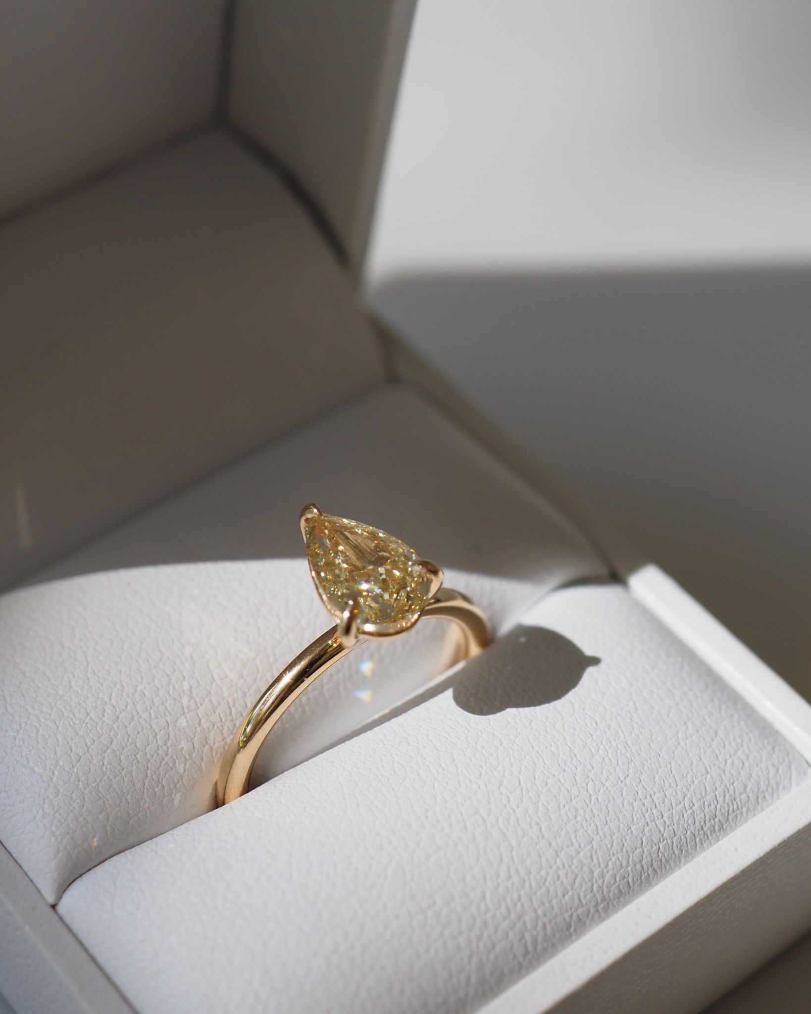 Gold ring with a pear-shaped yellow diamond set in an elegant band, showcased in a white jewellery box, bathed in soft light.