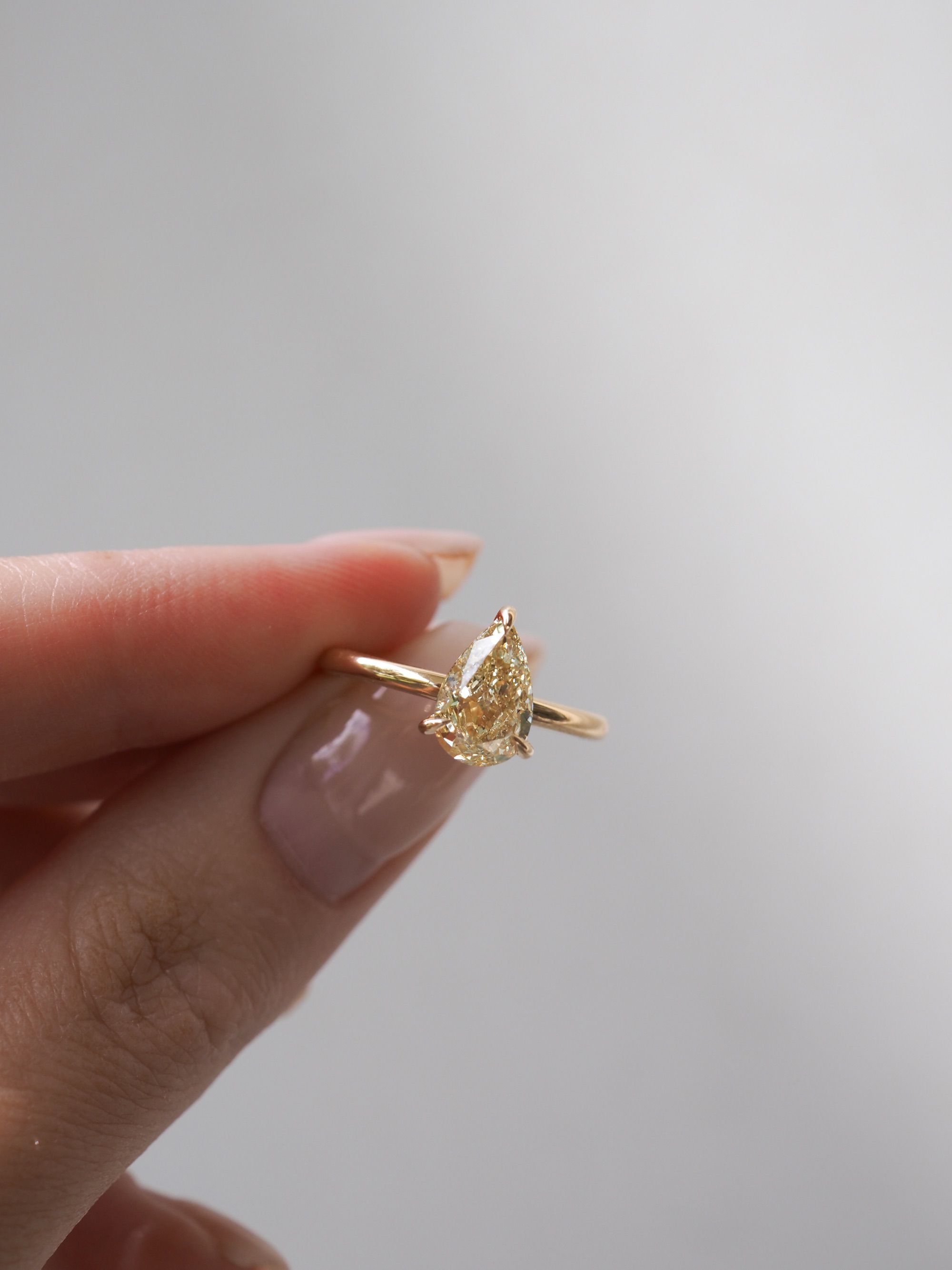 Hand holding an elegant marquise-cut yellow diamond ring with a gold band, against a soft grey background. Perfect luxury jewellery piece.