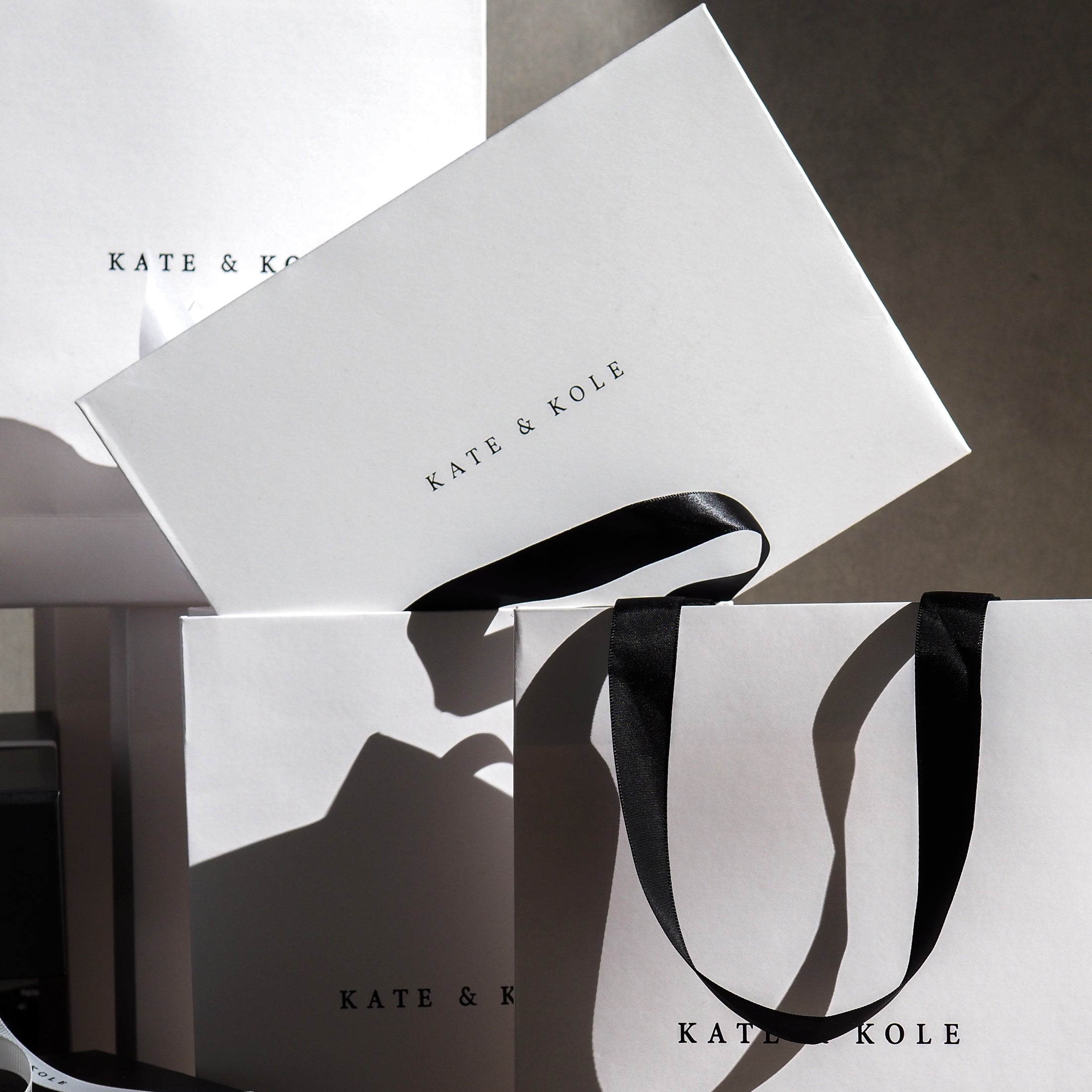 White "Kate & Kole" bags and boxes with black ribbon handles, elegantly arranged and bathed in soft light, suggest luxury jewellery.